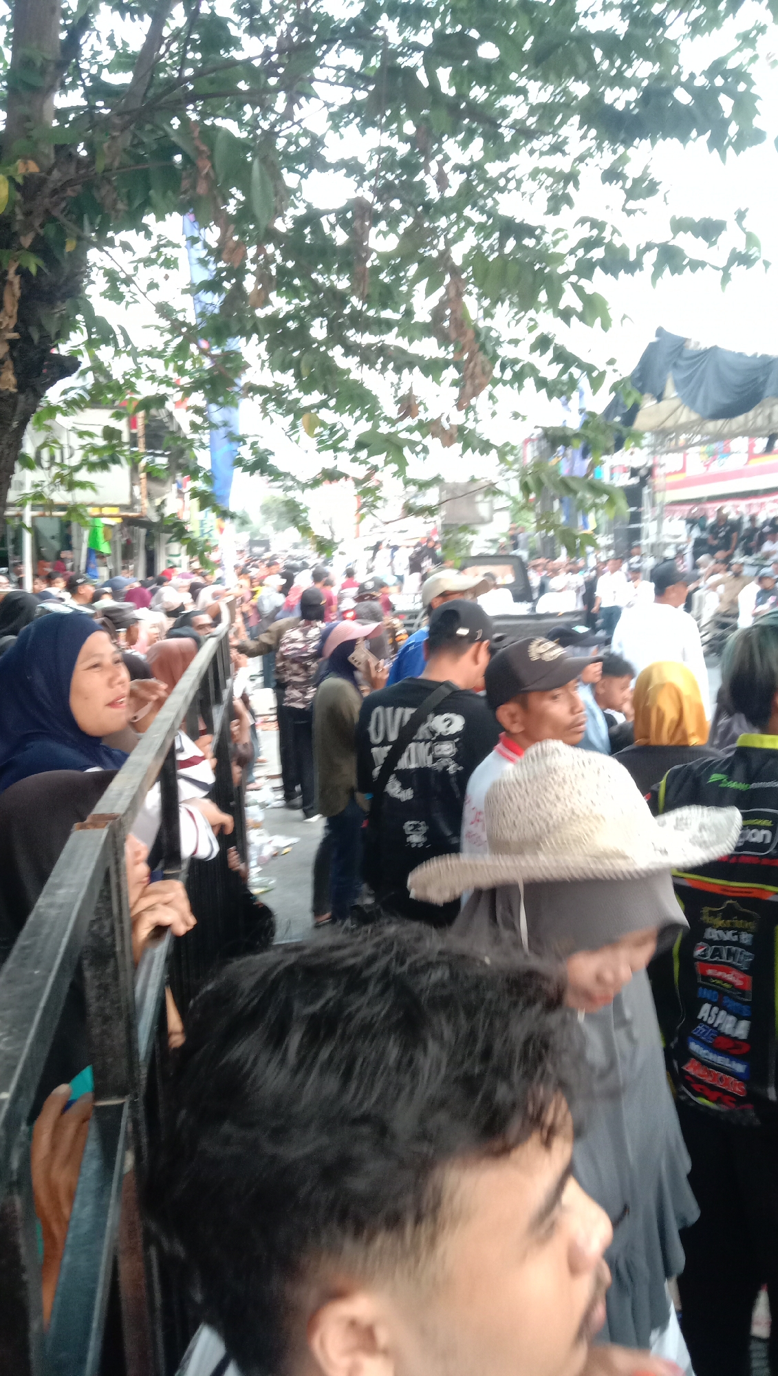 In Jatisawit, Indonesia, a crowd of people are gathered in what appears to be a street event, possibly a parade or festival. The image captures a section of the crowd from a slightly elevated perspective, looking over a metal barrier in the foreground. The people are diverse in age and attire, with many wearing casual clothing. Some individuals are seen wearing headscarves, indicating a blend of cultural influences.
In the foreground, a young man with dark, short hair is the most prominent figure. He is looking to his right, with his profile visible. Behind him and to his left, a woman wearing a dark blue hijab is looking upwards and to her right, with a relaxed expression. She is leaning against the metal barrier.
Further into the scene, a multitude of people are visible, some standing, others appearing to be moving. A few individuals are wearing hats, including a straw hat on a person in the middle ground. The background shows a street lined with buildings and stalls, suggesting a commercial or public area. Above the crowd, trees with green and dried leaves create a partial canopy, and there are hints of tents and structures, possibly for vendors or a stage. The overall impression is of a lively, outdoor gathering in a populated area.