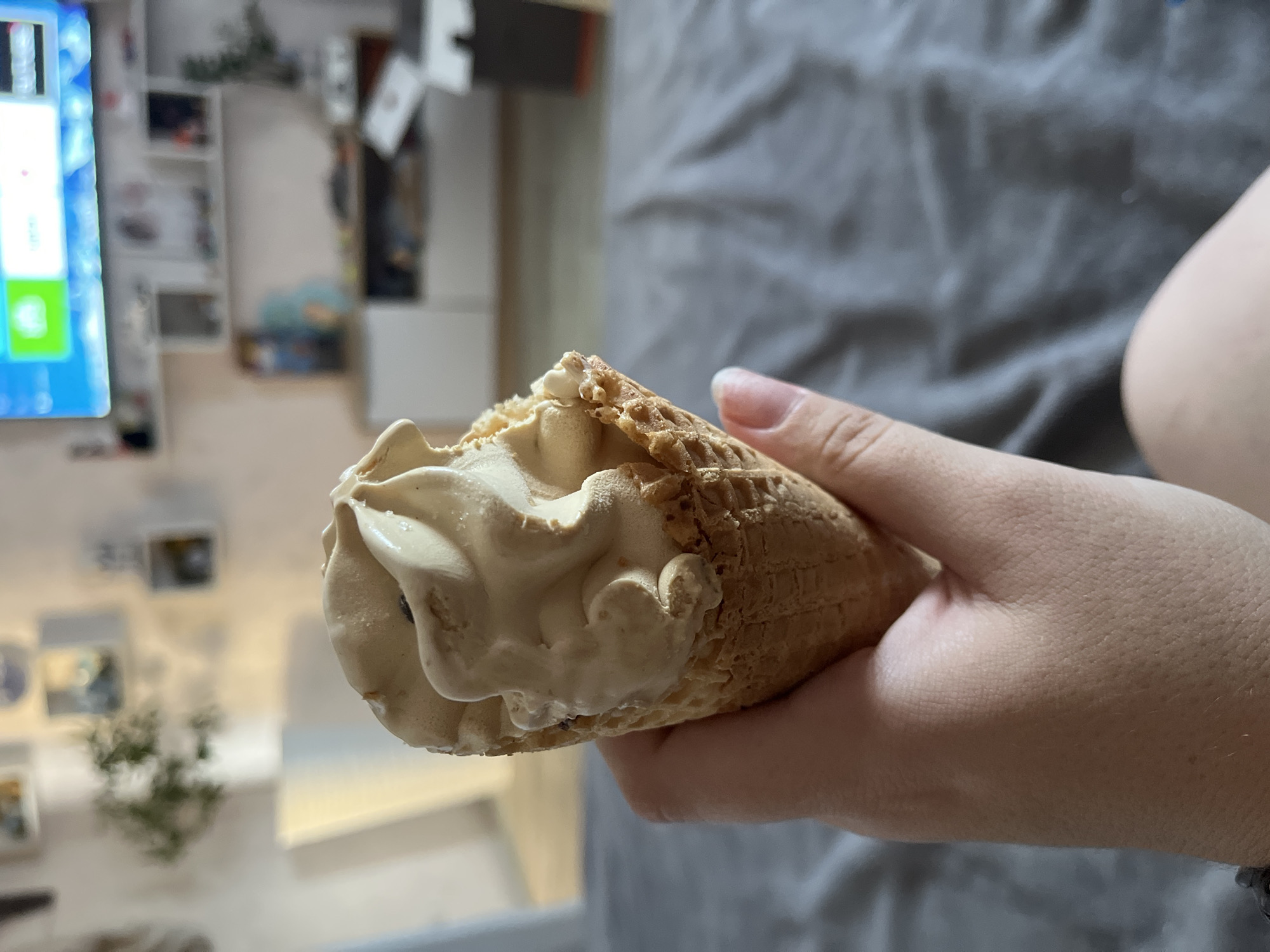 A close-up, shallow depth of field shot shows a person's hand holding a coffee-flavored ice cream cone. The ice cream is swirled in a waffle cone, with a bite taken out of it. In the background, out of focus, is a living room with a television on a stand, shelves with decorative items, and a plant. The scene takes place indoors, likely in a home. The location is not specified in the image.