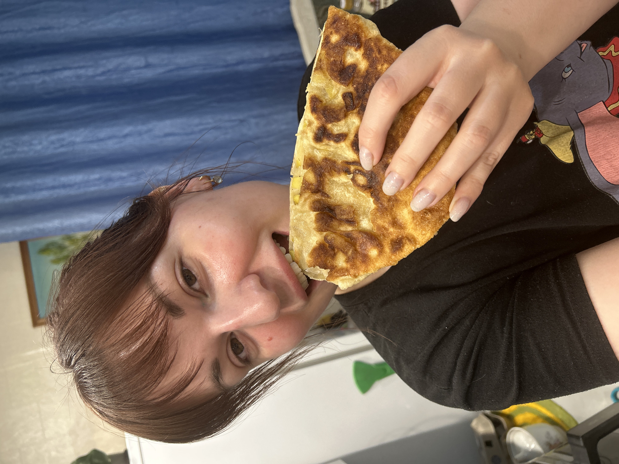 A woman is holding a large, triangular piece of food and taking a bite. The food appears to be a type of flatbread with a browned surface. She is wearing a black shirt. In the background, there is a blue curtain. The location cannot be confirmed using the image alone.