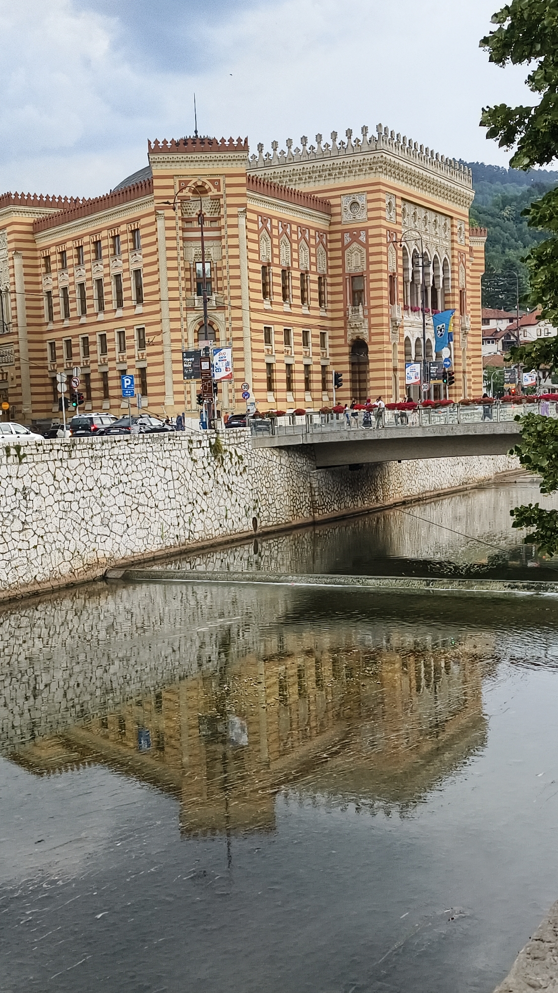 The image shows a view of the Miljacka River in Sarajevo, Bosnia and Herzegovina, with the Sarajevo City Hall (Vijećnica) reflected in the water. The City Hall is a grand, ornate building with striped yellow and red facades and intricate architectural details. It is situated on the bank of the river, with a stone embankment leading down to the water. A bridge spans the river in front of the building, with people walking across it and cars on the road. The reflection of the City Hall in the water is clear, though slightly distorted by the rippling surface of the river. Trees and a hilly landscape can be seen in the background. The sky is overcast, suggesting a cloudy day.