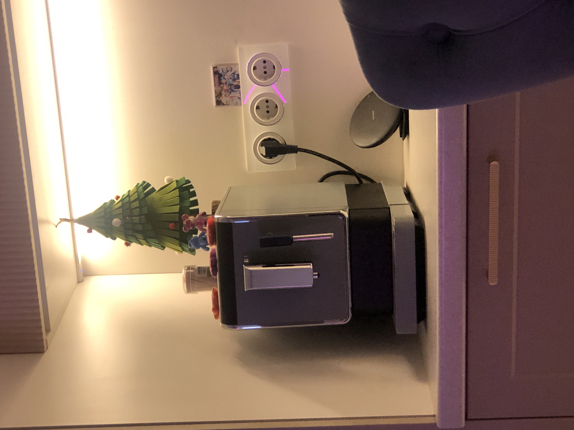 This image depicts an indoor scene, likely a dedicated coffee or beverage station, situated in Saint Petersburg, Russia. A modern dark grey and chrome coffee machine occupies the center of a light-colored countertop. Above the coffee machine, a green, multi-layered felt Christmas tree decoration, adorned with white pom-poms and red accents, hangs from an unseen fixture.
On top of the coffee machine, a clear plastic container with a light lid stands behind two small figures: one blue and one pink, both resembling cartoon bears. Several small, reddish-purple objects are also scattered on the machine's surface.
To the right, a white triple electrical outlet strip is mounted on a light-colored wall. A black power cord extends from the coffee machine and is plugged into the leftmost Type F (Schuko) socket. The rightmost socket is illuminated by a distinct pink-purple light pattern. The central socket remains empty. Above the outlets, a small rectangular picture or magnet displays an image of multiple cartoon-style figures or children.
Further right on the countertop, a black circular wireless charging pad is positioned. The entire area is brightly illuminated by a warm yellow light source, likely an LED strip, located beneath an overhead shelf or cabinet. A dark blue upholstered object, consistent with an armrest or backrest of a chair, is partially visible in the lower right foreground. Below the countertop, a light purple-grey cabinet with a horizontal, textured handle is visible. No individuals are directly visible in the frame.