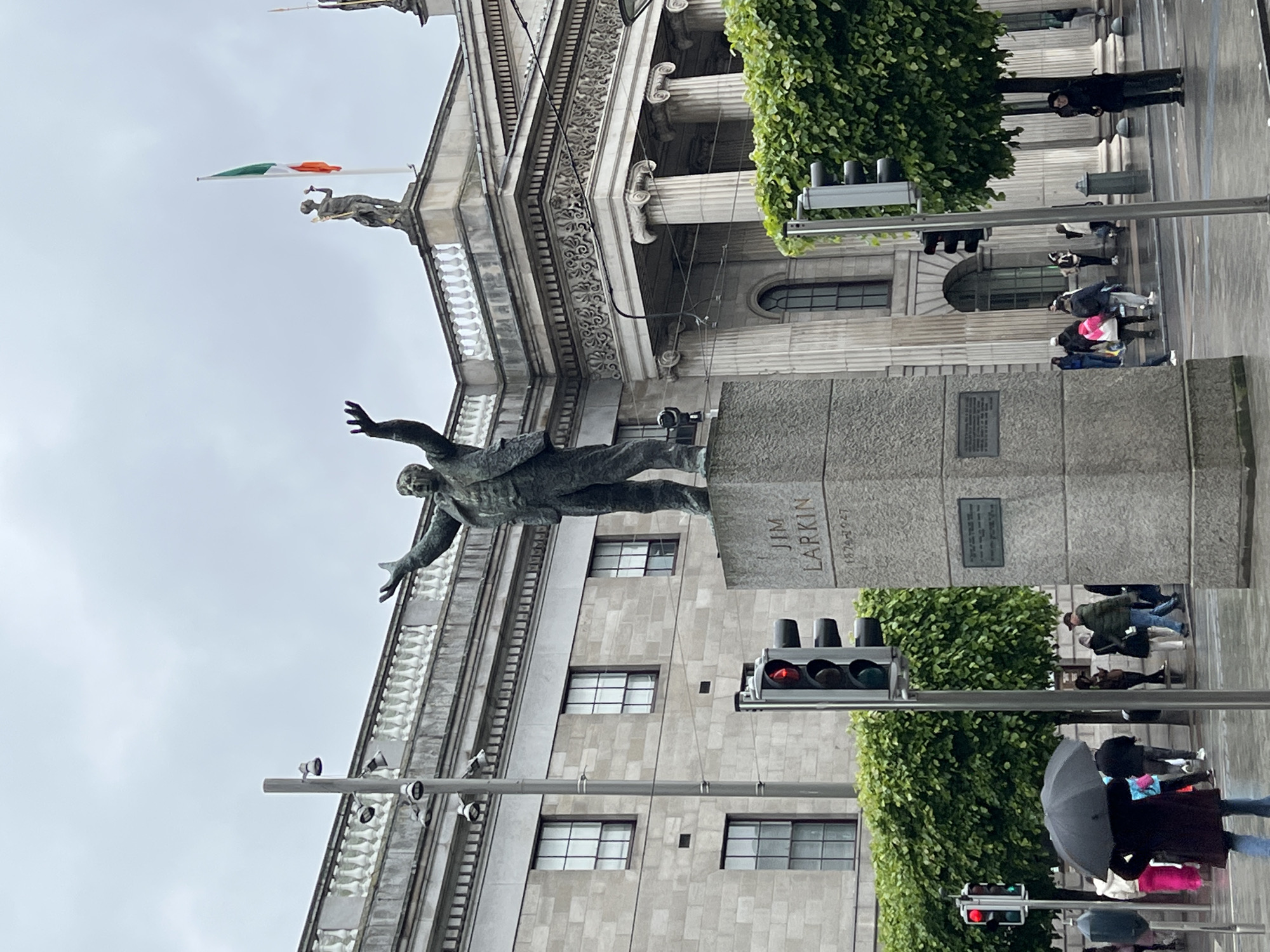 This image shows the Jim Larkin statue in O'Connell Street, Dublin, Ireland. The statue depicts a man with arms outstretched, mounted on a large stone plinth. A building with columns and an Irish flag can be seen behind the statue. Pedestrians are walking along the street, some carrying umbrellas. Traffic lights are visible, indicating the presence of a street intersection. Green foliage is present on the side of the building. The sky is overcast.