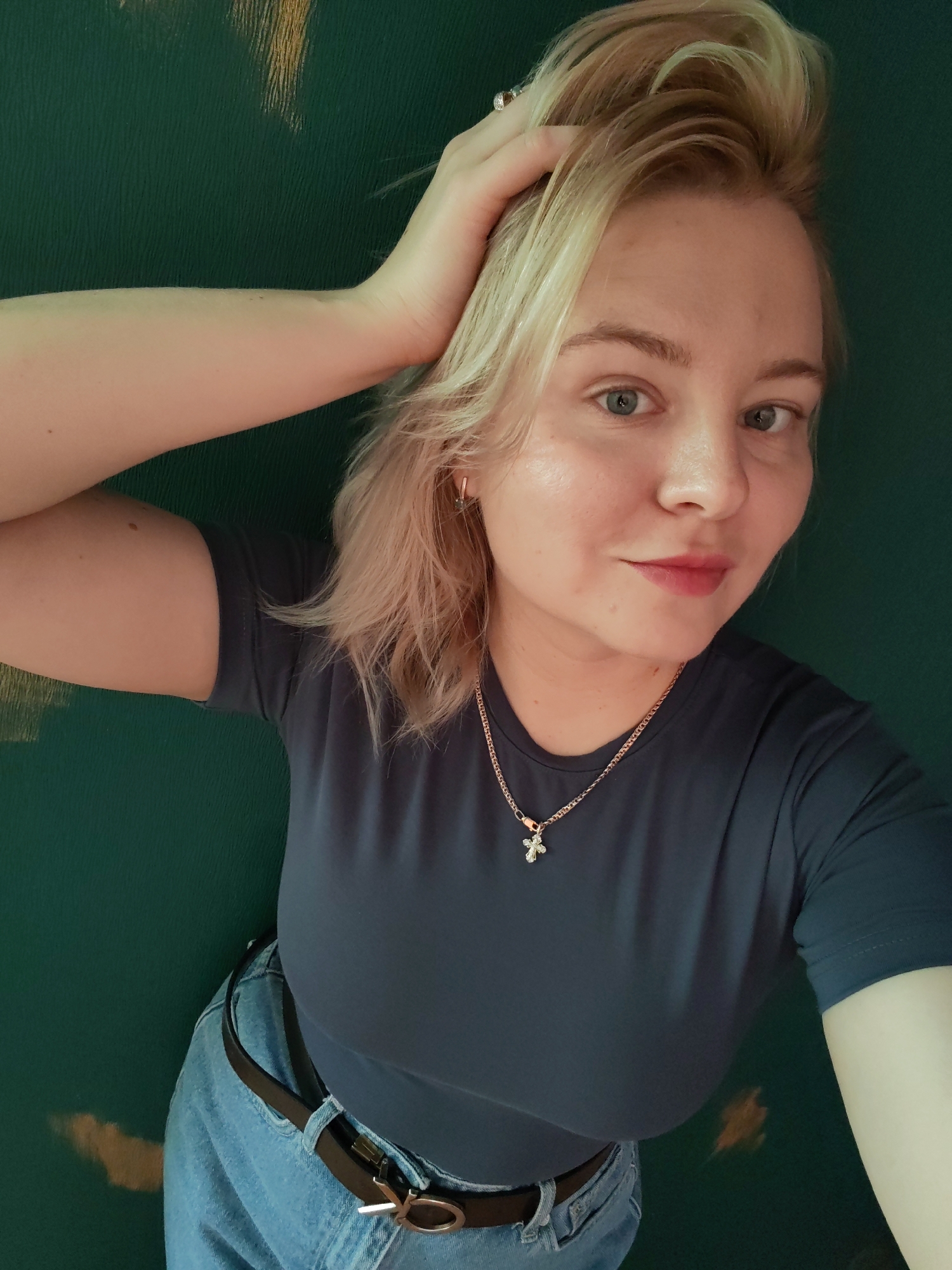 A blonde woman is posing for a selfie against a textured green wall. She has her right hand raised, touching her hair, and is wearing a dark blue t-shirt, blue jeans, and a black belt with a silver buckle. A rose gold necklace with a cross pendant is visible around her neck. The image does not contain any information about the location in Izhevsk, Russia.