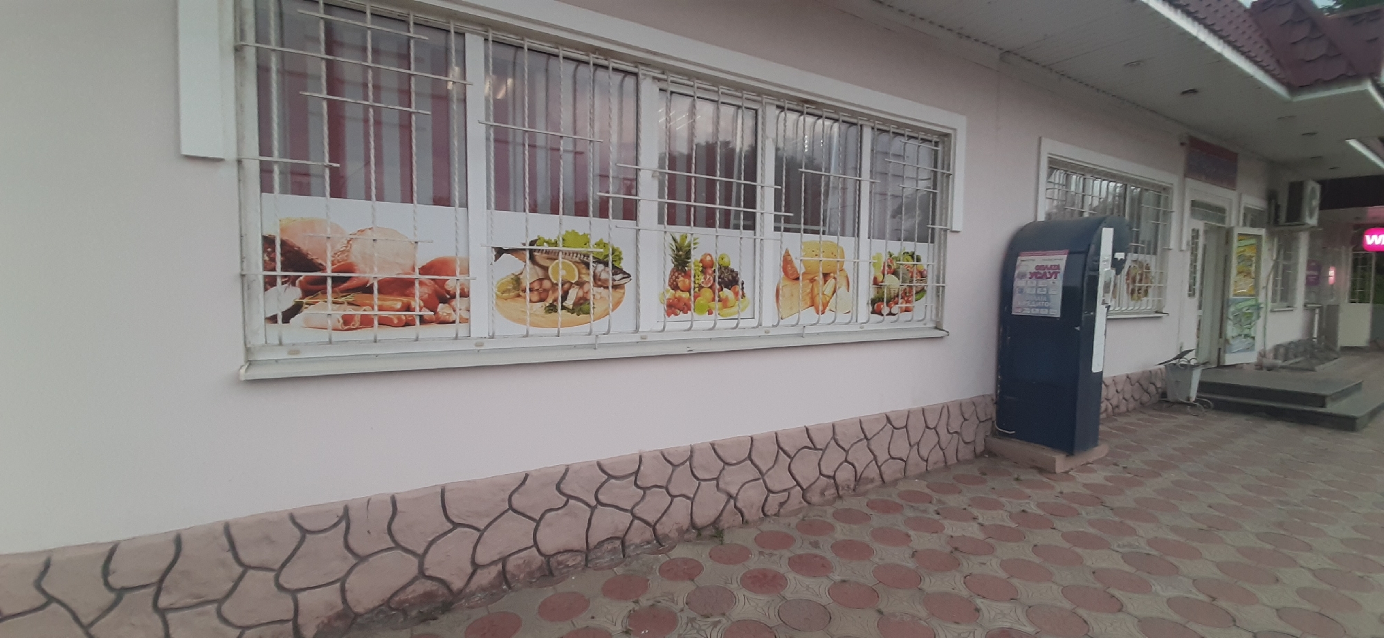 The image shows the exterior of a building in Yessentukskaya, Russia, likely a small shop or market. The building has a light pink facade and is equipped with barred windows. These windows display large photographic posters of food items, including meat products, fish, fruits, and bread. In front of the building, there is a dark blue kiosk, possibly a payment terminal or information booth, with Cyrillic text on its side. The ground in front of the building is paved with reddish-brown interlocking bricks. A tiled base runs along the bottom of the building wall. The overall scene is static, with no people visible.