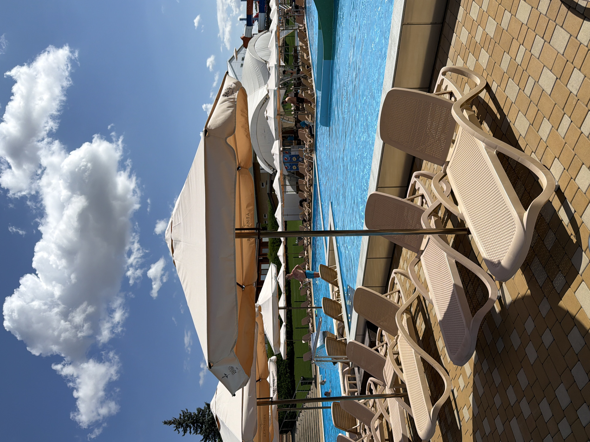 The image depicts a sunny day at a swimming pool complex, likely in Kamyshin, Russia, given the prompt. In the foreground, rows of empty beige lounge chairs are neatly arranged on a paved patio made of brown and beige interlocking bricks. Several large, cream-colored umbrellas provide shade over some of the chairs.
Beyond the lounge chairs, a bright blue swimming pool dominates the middle ground. The water is clear, and several people are visible in the pool, some swimming and others standing. One man in blue swim trunks is standing at the edge of the pool, facing away from the camera. In the background, more lounge chairs and umbrellas are set up, along with a grassy area and buildings. To the right, a section of a water slide with red and white elements is visible. The sky is a vibrant blue with large, fluffy white clouds, and a tall evergreen tree stands to the far left of the frame. The overall scene suggests a recreational area designed for relaxation and enjoyment on a warm day.