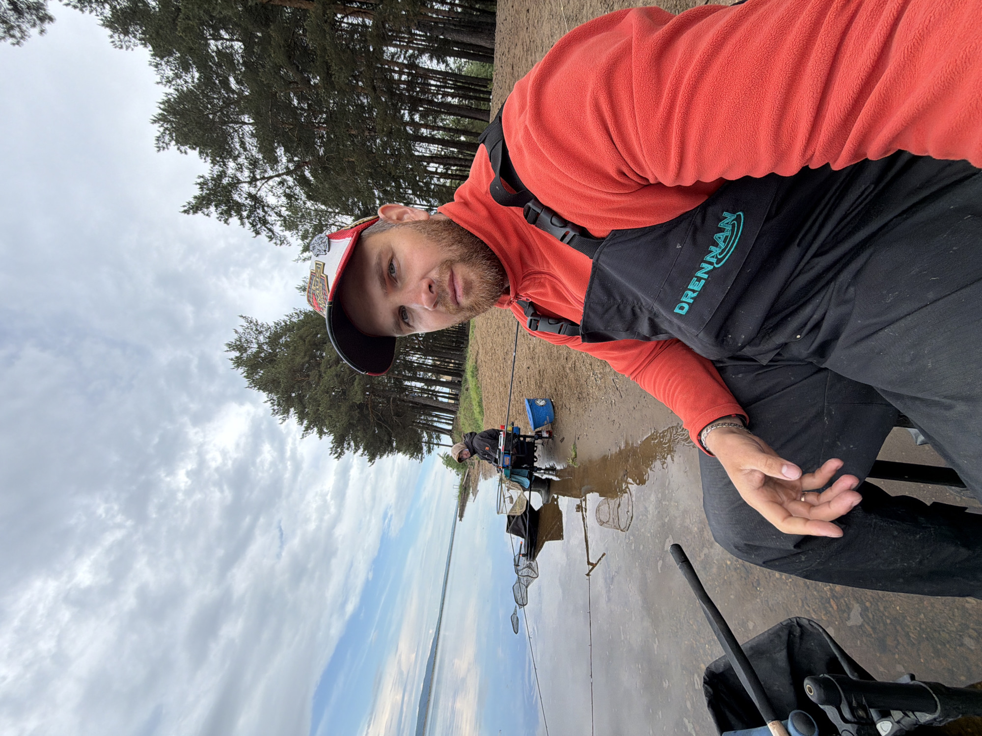 A medium-shot, outdoor photo shows a man in the foreground, appearing to take a selfie, with a lake and shoreline visible in the background. The location is likely Chaikovsky, Russia, given the prompt.
The man is wearing an orange fleece, black bib overalls with "Drennan" printed on them, and a baseball cap. He has a beard and is looking towards the camera. His right hand is visible, showing a silver bracelet and a wedding band on his ring finger.
Behind the man is a muddy shoreline transitioning into a large body of water, presumably a lake. In the distance, another person is seated on a fishing platform, equipped with various fishing gear. Several fishing nets are visible, extending into the water. The sky is overcast with a mix of clouds and blue patches. Trees line the shoreline in the distance. Additional fishing equipment is visible near the man in the foreground, and appears to be on or near his chair.