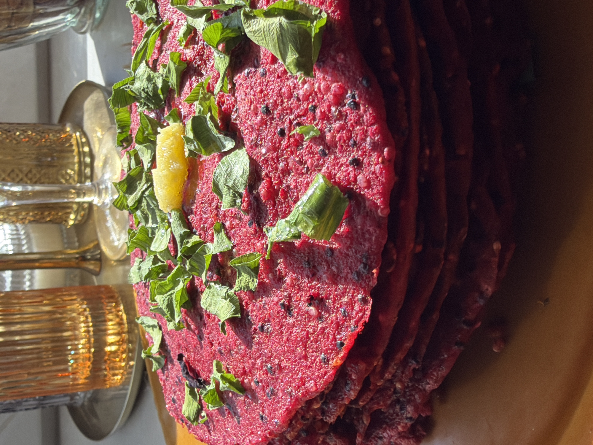 The image shows a stack of red flatbreads, possibly beetroot flatbreads, garnished with green leaves and a yellow slice, possibly a citrus fruit, on a light brown surface. To the left of the flatbreads, there is a blurry view of ornate glassware, possibly metal-rimmed, with a gilded appearance. The lighting suggests an indoor setting with natural light coming from the left. There is no information about the location in Krasnoyarsk, Russia in the image.