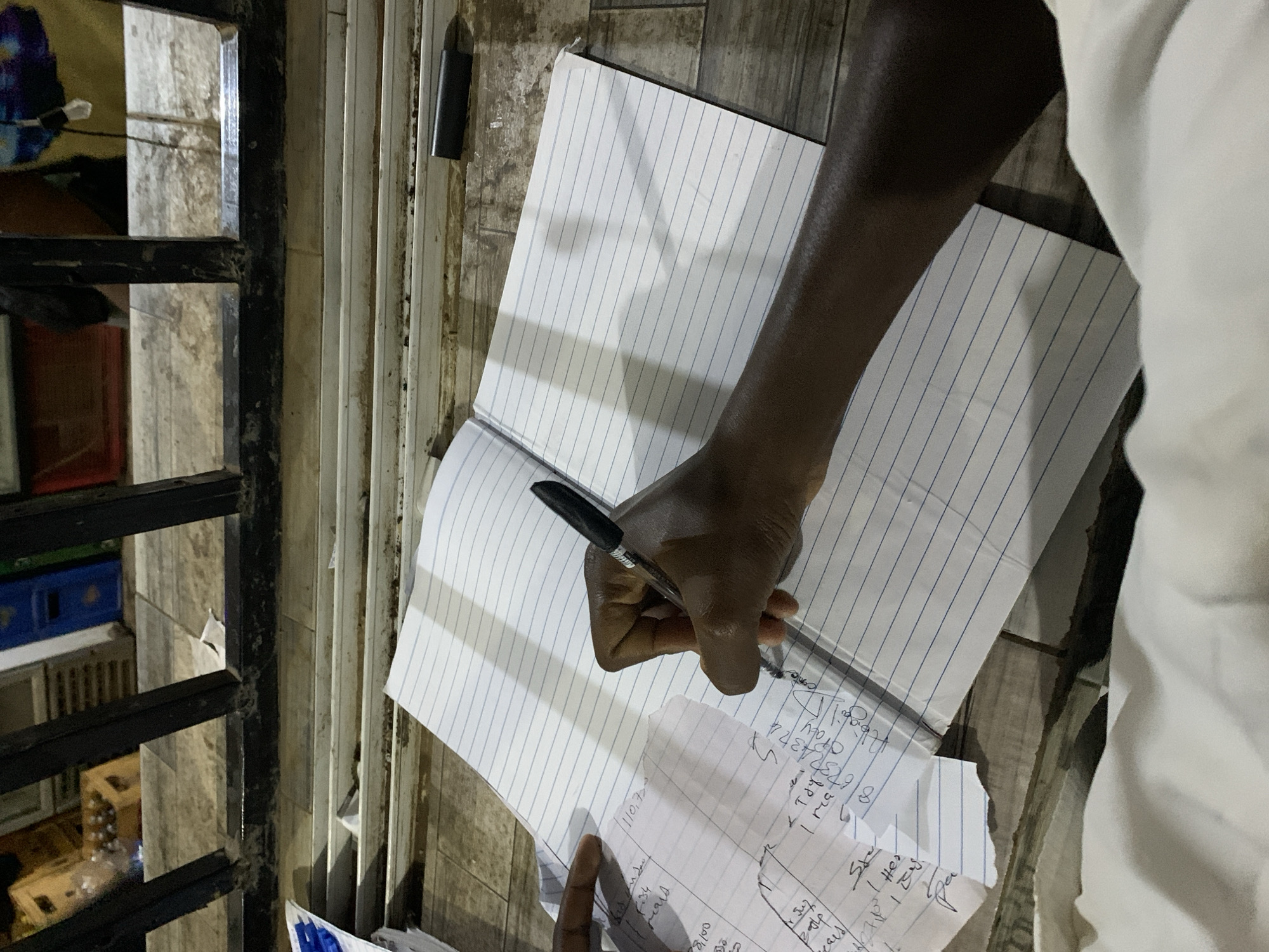 The scene is observed in New Stadium, Nigeria. A dark-skinned individual's right arm and hand are visible from an overhead perspective, actively writing on a white lined notebook with a black ballpoint pen. A torn piece of white lined paper containing handwritten numerical and textual data is positioned to the left of the notebook, partially secured by a visible left hand, indicating active reference. The writing surface consists of light-colored, worn wooden or tiled planks showing dirt and discoloration. In the background, a dark, weathered metal railing with vertical and horizontal bars delineates the immediate area from a storage or commercial space. Beyond the railing, brown wooden crates holding bottles are visible on the left, and blue plastic crates are present on the right. A small, dark rectangular object, possibly an eraser or pen cap, rests on the wooden surface to the right of the notebook. A section of white fabric is visible at the bottom of the frame.