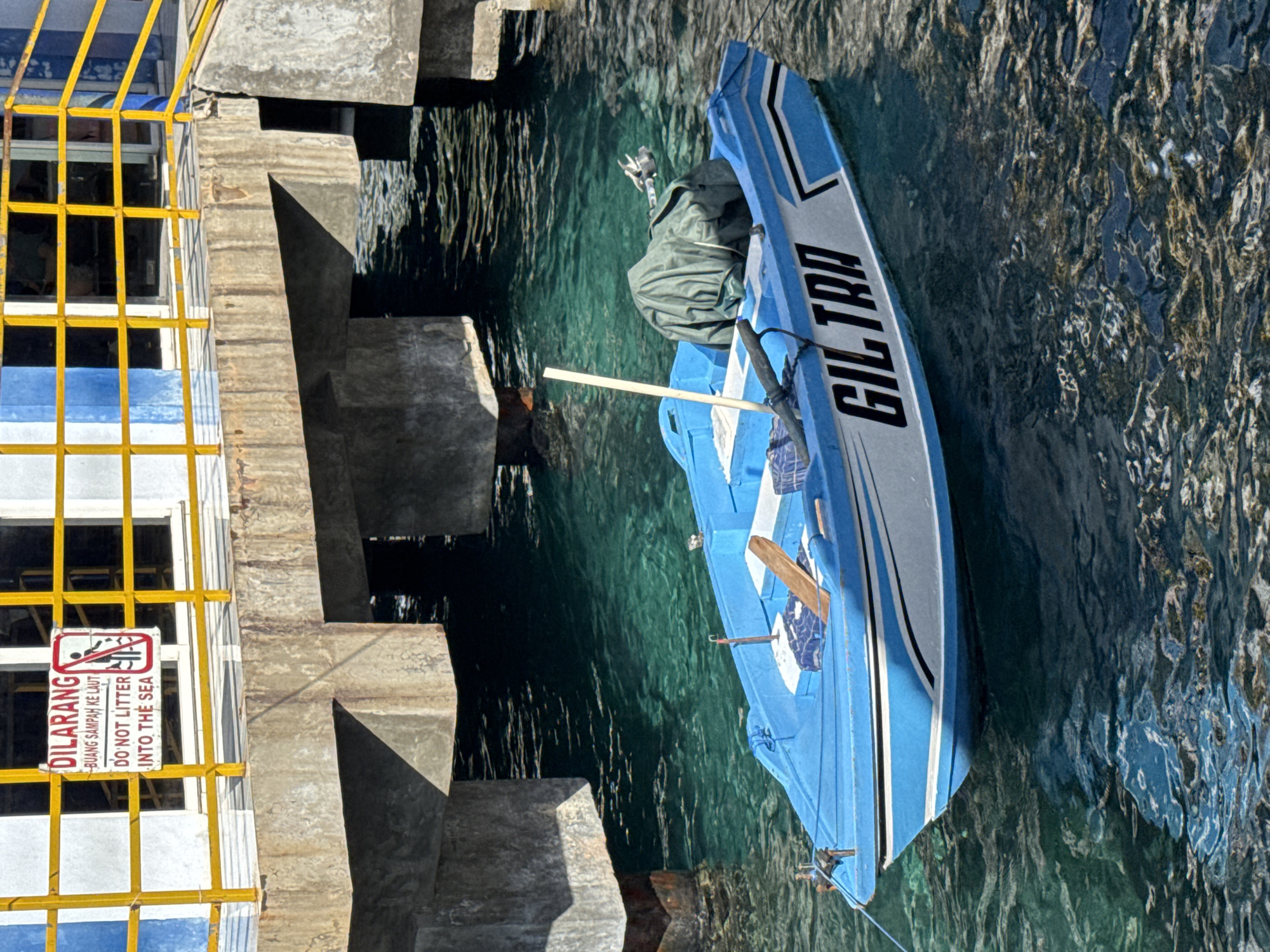 A small blue and white boat, named "GILTRA" in prominent black lettering, is moored alongside a concrete pier under bright sunlight. The boat, appearing to be a local transport or fishing vessel, features a covered engine or equipment under a green tarp and a wooden paddle. The water is clear, calm, and a striking turquoise-green, reflecting ripples on its surface.
The concrete pier, made of large blocks, runs along the left side of the frame. Above the pier, a bright yellow metal railing, part of a larger structure, is visible. Attached to this railing is a white warning sign with a red border, depicting a crossed-out person littering. The sign bears the text "DILARANG BUANG SAMPAH KE LAUT" (meaning "Forbidden to throw trash into the sea") in Indonesian, followed by the English translation: "DO NOT LITTER INTO THE SEA."
Strong, clear shadows indicate that the image was captured during the day, likely mid-morning or early afternoon. No individuals are present in the scene, which conveys a peaceful and functional atmosphere typical of a coastal dock in Indonesia.