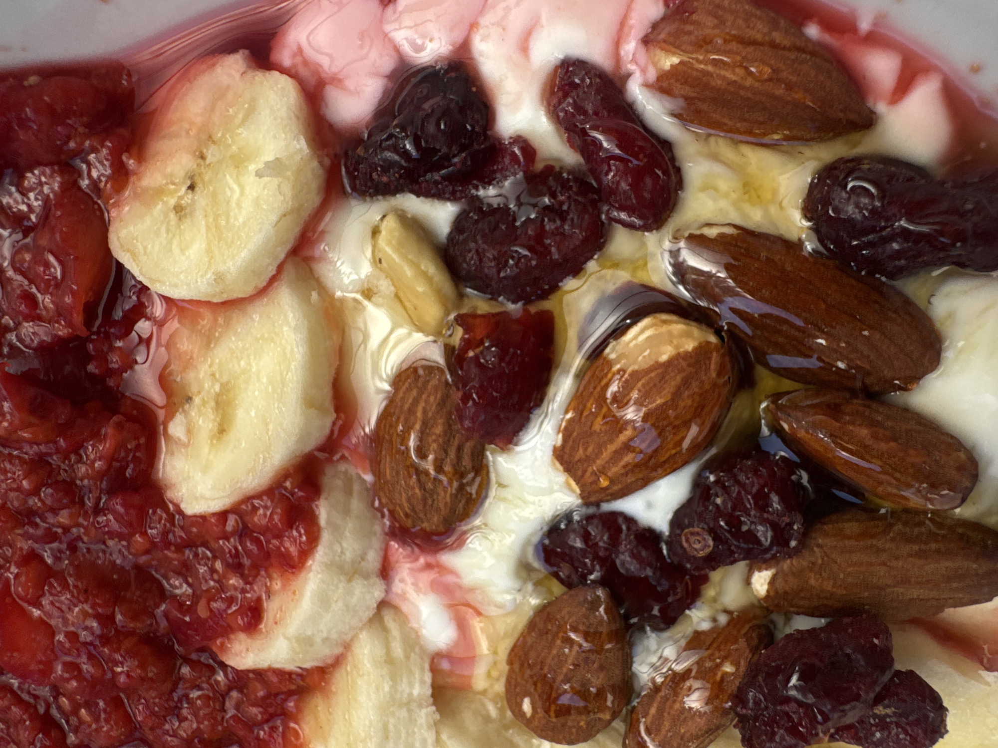 A close-up, overhead shot shows a bowl of yogurt topped with raspberries, banana slices, almonds, and cranberries. The yogurt is a creamy white, with streaks of pink and red from the fruit and perhaps a berry sauce. The raspberries are mashed and form a vibrant red layer on top, interspersed with the pale yellow banana slices. The almonds are whole and brown, some appearing to be coated in honey or syrup, which also glistens on the cranberries, giving them a dark, glossy appearance. A few pale yellow nuts, possibly peanuts, are also visible amongst the other toppings. The bowl itself is not fully visible, but the arrangement of the ingredients suggests a healthy breakfast or snack. The lighting is bright, highlighting the textures and colors of the food.