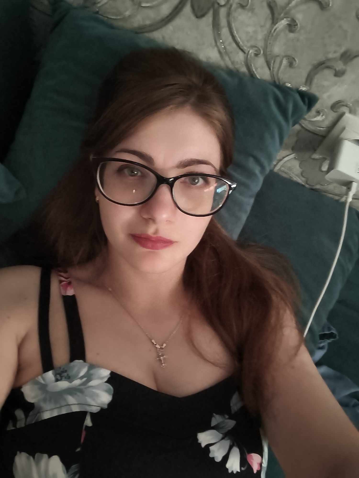 In a medium shot, a woman with long, brown hair and glasses is lying down, looking at the camera. She is wearing a black dress with a floral pattern and a necklace with a cross pendant. She is wearing light pink lipstick and has green eyes. She is on a teal-colored pillow, which is on a bed with a patterned headboard. There is a white electrical outlet visible on the right side of the image. The scene appears to be indoors, possibly in a bedroom. There is no indication of the specific location within the city of Novaya Usman', Russia.