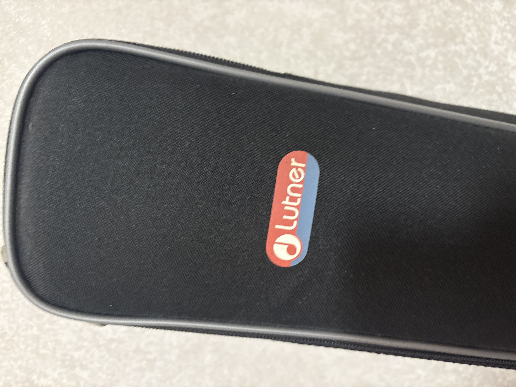 A black, rectangular case with a gray zipper and trim sits on a textured, light-colored surface. A red and blue oval-shaped logo with the text "Lutner" in white is affixed to the front of the case. The logo features a stylized musical note. The surface appears to be a countertop or table. The scene is static and shows no movement or interaction. The image does not provide any information about the city of Kazan, Russia.