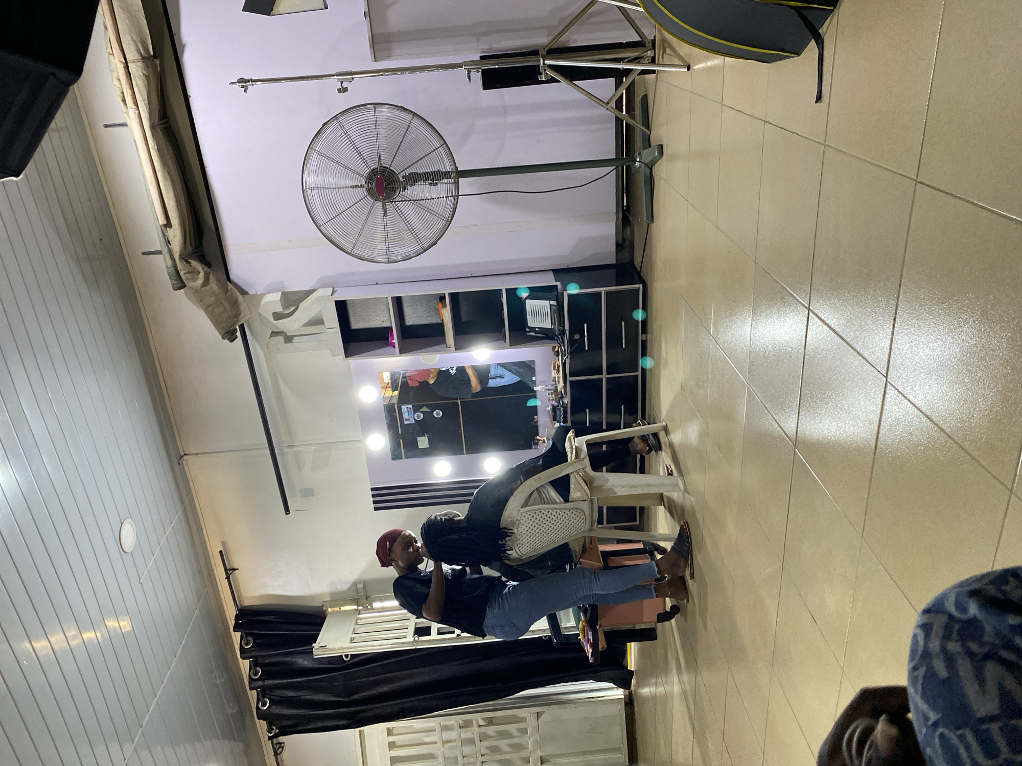 The image depicts an interior scene, likely a beauty salon or hair styling studio. A woman with a red headscarf, wearing a black t-shirt and blue jeans, is standing next to a seated individual. The woman appears to be styling the hair of the seated person. Behind them is a lighted vanity mirror, surrounded by black shelving. A large fan is mounted on the wall near the vanity, and other equipment is seen in the room. The flooring consists of light-colored tiles. The location is indicated as Bukuru, Nigeria.