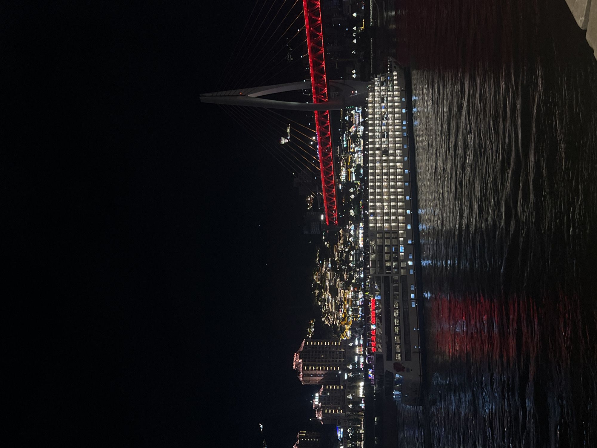 A long cruise ship is illuminated by countless small windows as it sails on dark water at night. The ship is positioned in the foreground, occupying the lower half of the frame. Behind the ship, a brightly lit cityscape is visible, with many buildings showcasing illuminated windows and decorative lights. A large, modern bridge with red lights spanning its length stretches across the scene in the middle ground, with a prominent white tower supporting its structure. The night sky above is dark and clear, with a few faint specks of light visible. The water reflects the red lights of the bridge and the white lights of the ship, creating a shimmering effect. The scene appears to be a riverfront area in Yuzhong District, China, characterized by its urban development and nighttime illumination.