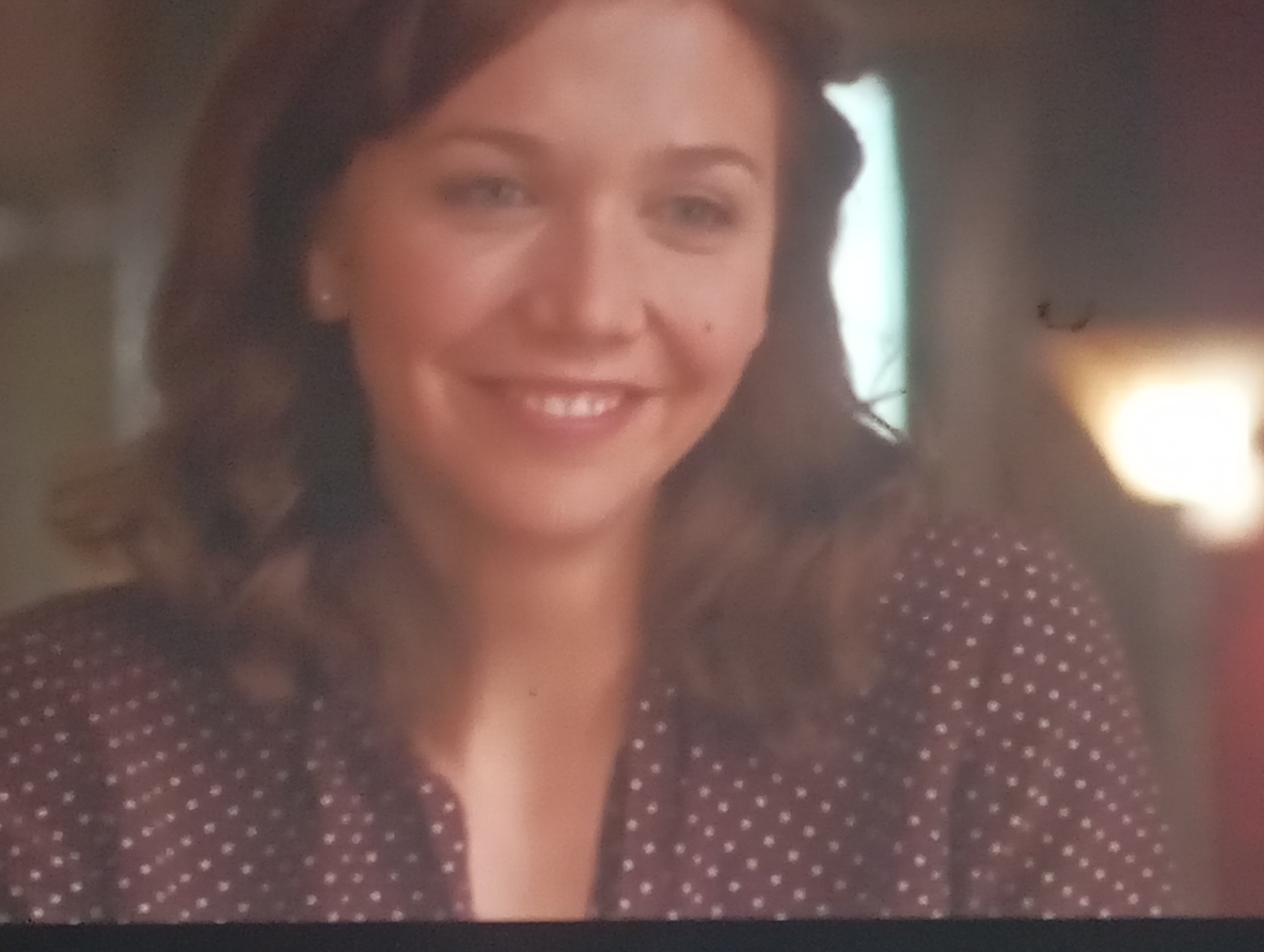 This is a close-up, medium-shot media file featuring a woman. She has fair skin, light brown hair, and is looking towards the viewer with a slight smile. She is wearing a dark, possibly burgundy, shirt with a white polka dot pattern. The background is softly lit and appears to be indoors, with hints of a bright light source to the right, possibly a lamp, and a cooler light to the left, which might be a window. The overall mood is warm and inviting. There are no visible text elements. The specific location within Sofiivska Borshchahivka, Ukraine, cannot be determined from the visual information provided.