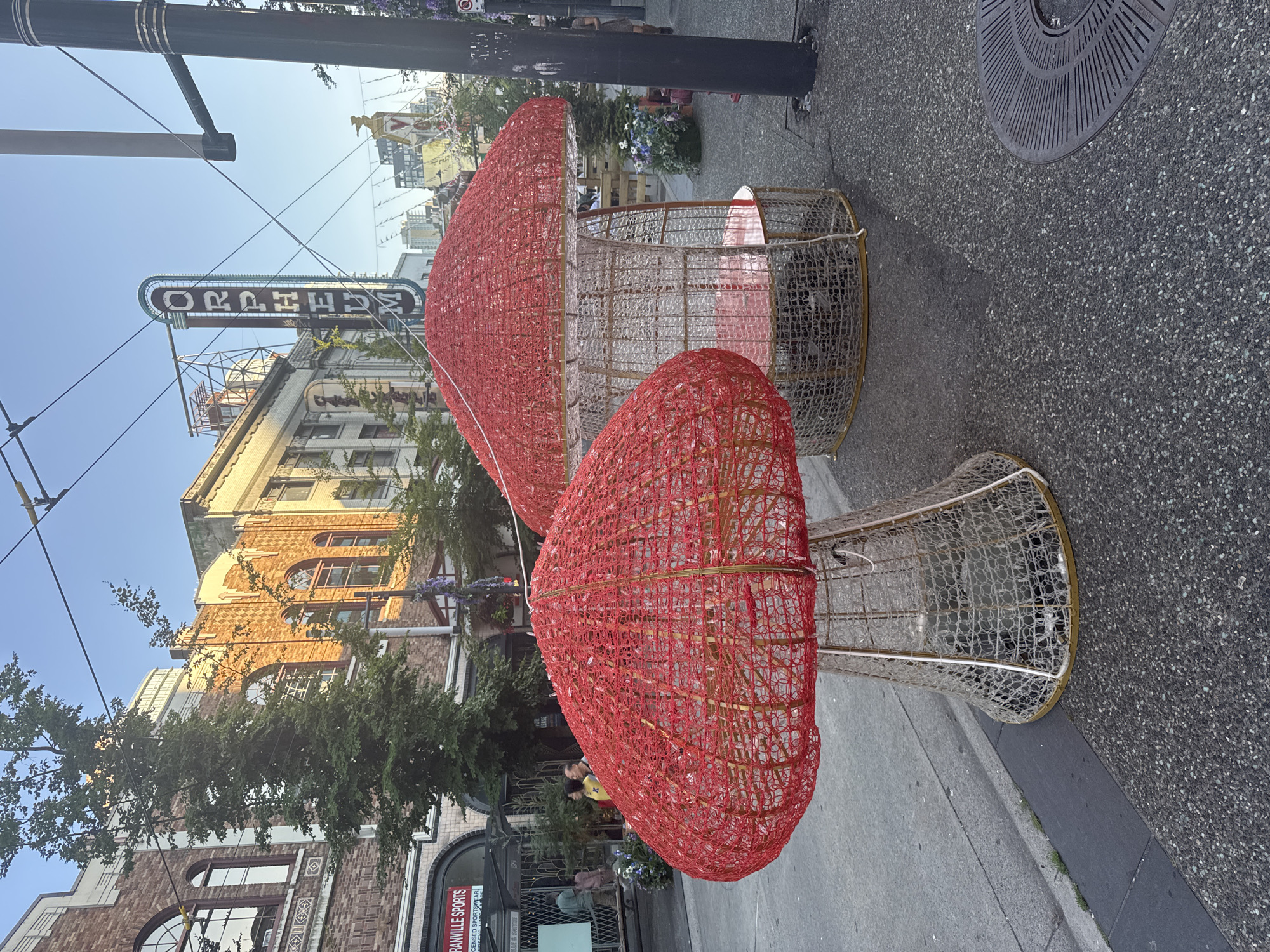 The image captures a street scene in downtown Vancouver, Canada, likely in the vicinity of the Orpheum Theatre, given the prominent "ORPHEUM" marquee visible in the background. Two large, decorative mushroom-shaped structures, illuminated with red mesh or lights, are the primary subjects in the foreground. The structure on the left is closer and appears to be a solid mushroom cap atop a woven base. The structure on the right is larger and open, resembling a small pavilion or seating area, with a red cap and a woven cage-like structure containing a pink seat.
The street is paved with asphalt. To the left of the mushrooms, a sidewalk is visible, with a curb separating it from the street. Behind the mushrooms, on the left side of the image, is the exterior of a building with brickwork and arched windows, featuring a sign for "GRANVILLE SPORTS" and "LICENSED SPORTSWEAR." Several people are present on the sidewalk in front of this building, appearing to be patrons or pedestrians.
The background reveals more of the urban environment, including more buildings with intricate architectural details, trees, and overhead wires, typical of a city street. The sky is clear and blue, suggesting a daytime scene, possibly late afternoon due to the warm lighting on the buildings. A lamppost is visible on the right side of the image. The overall impression is of a vibrant, decorated streetscape in an urban setting.
