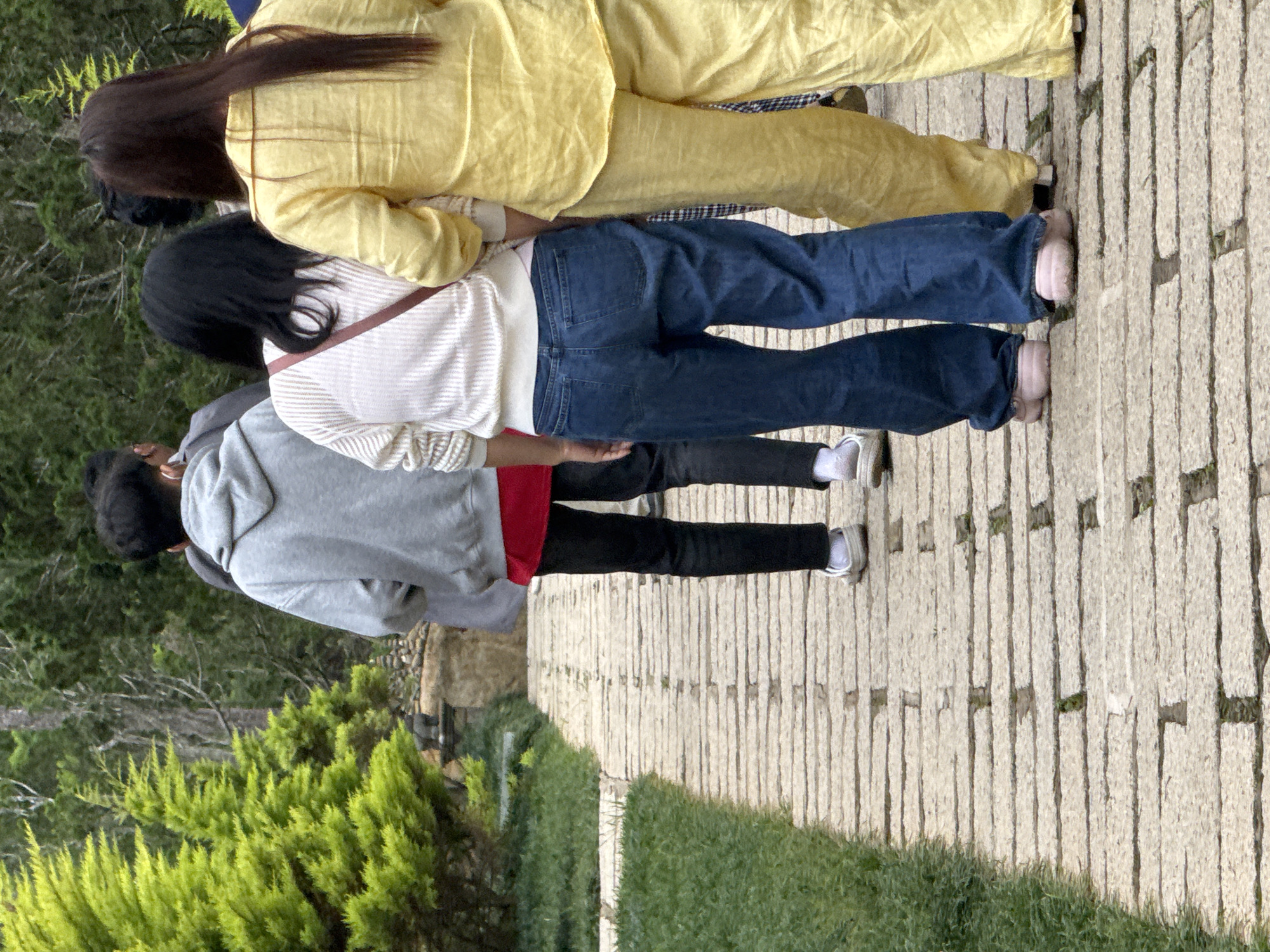 In Da Lat, Vietnam, four individuals walk along a stone pathway lined with lush green grass and trees. The first person from the left, partially visible, wears a grey hooded sweatshirt over a red shirt and black pants. Next, a person in a white, long-sleeved shirt and blue jeans walks ahead, with a small bag strapped across their chest. To their right, a person in a bright yellow ensemble, likely a coordinated top and pants set, follows. The fourth individual, on the far right, is largely obscured, with only a portion of their leg visible. The path is constructed with light-colored rectangular stones, and the surrounding vegetation suggests a park or garden setting.