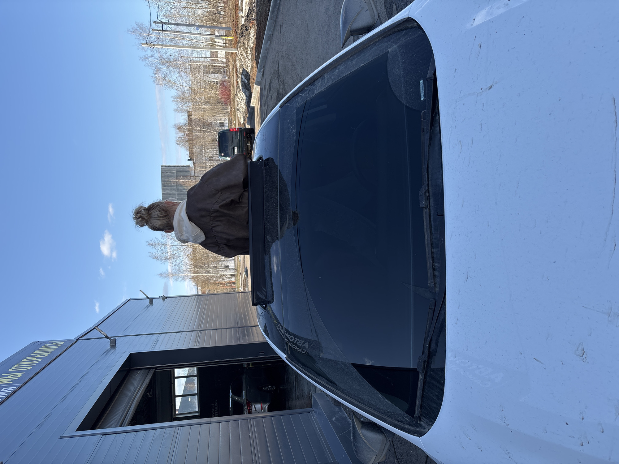 A medium, eye-level shot depicts the front of a white, dirty vehicle, positioned at an angle. The vehicle is situated in front of a gray building, part of which is visible on the left side of the frame. Above the building, the sky is a clear, light blue. A female figure is visible, leaning into the vehicle through the open roof. She is wearing a brown jacket over a white garment, with her blonde hair pulled back in a bun.
In the background, there are trees and some buildings, indicating an urban setting. The text on the building on the left side of the image is Russian. Based on this, and the style of the buildings, it is very likely the location is Izhevsk, Russia. A dark-colored vehicle is parked behind the white one. The image appears to be taken on a sunny day with a clear sky.