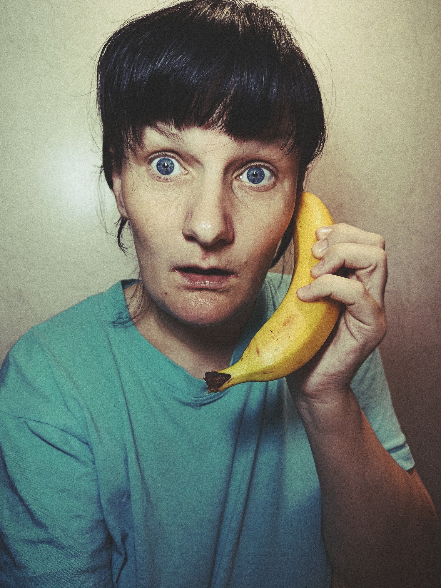 A woman with short dark hair and wide blue eyes holds a banana up to her ear as if using it as a telephone. She wears a turquoise t-shirt. The background is a textured beige wall. The location is not discernible from the image.