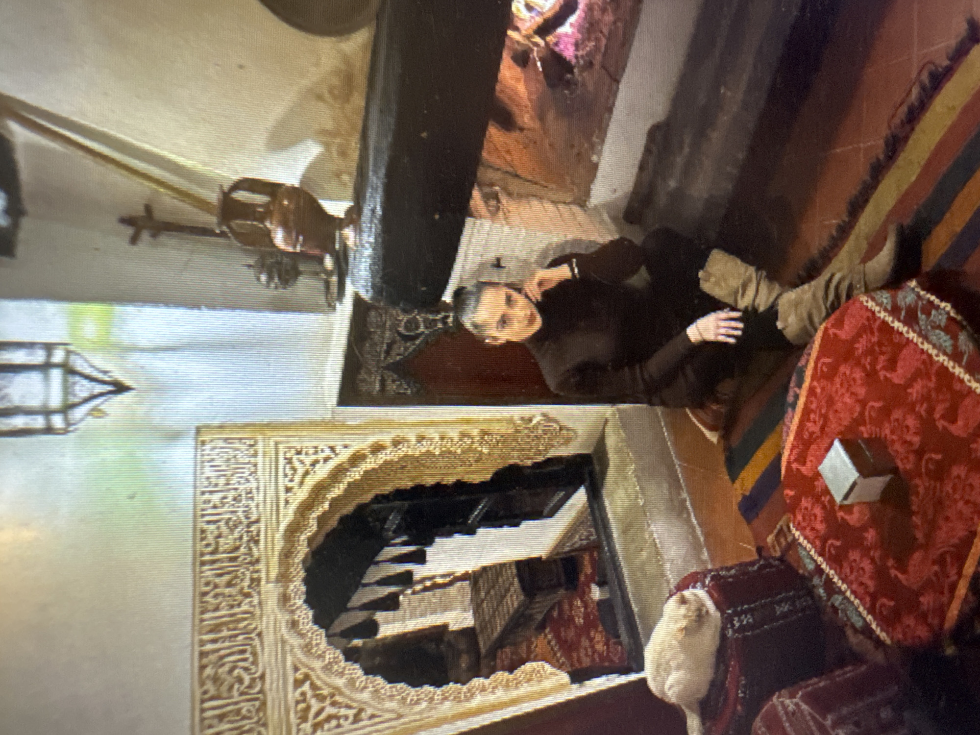 The image depicts an interior scene that appears to be a living room or lounge area with traditional Moroccan-inspired decor. In the foreground, a woman with dark hair pulled back in a bun is seated on the floor, leaning on her hand. She is wearing a dark, high-necked, long-sleeved top and tan, knee-high boots. In front of her is a low, red table with intricate patterns and fringe, topped with a small, cube-shaped object. To her left, a white cat is curled up on a red cushion. Behind the woman, a large, dark fireplace is visible, with a fire burning inside. To the left of the fireplace, an ornate arched doorway or alcove is present, decorated with intricate carvings and calligraphy. Through the arch, a glimpse of another room is visible, featuring a low, wooden bench or table and a red carpet. The walls are a light, textured color. The overall impression is one of a cozy, culturally rich environment. However, there are no discernible elements in the image that would allow for the identification of the city as Murcia, Spain.