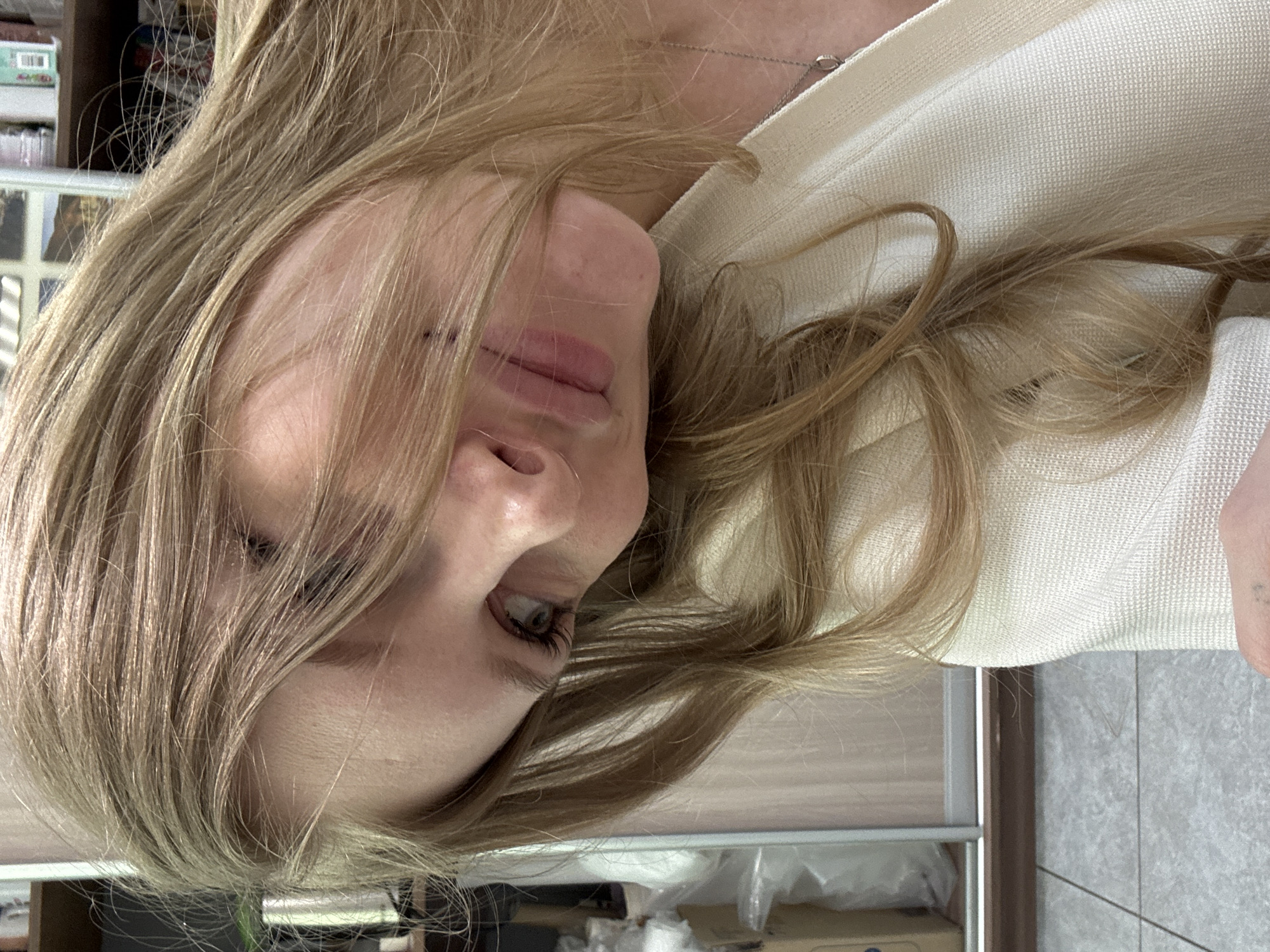 The image shows a close-up of a blonde woman's face. Her hair is light brown, parted on the left, and falls across her face. She has pale skin, a small nose, and pink lips. Her right eye is visible and appears to be hazel or green. She is wearing a light-colored, textured fabric garment, possibly a sweater or blouse. A silver necklace is visible around her neck. The background shows a partial view of a room with a light-colored cabinet or shelving unit, and a tiled floor. The location in the city of Perm, Russia is not discernible from the provided visual information. The image focuses primarily on the woman's face and upper body.
