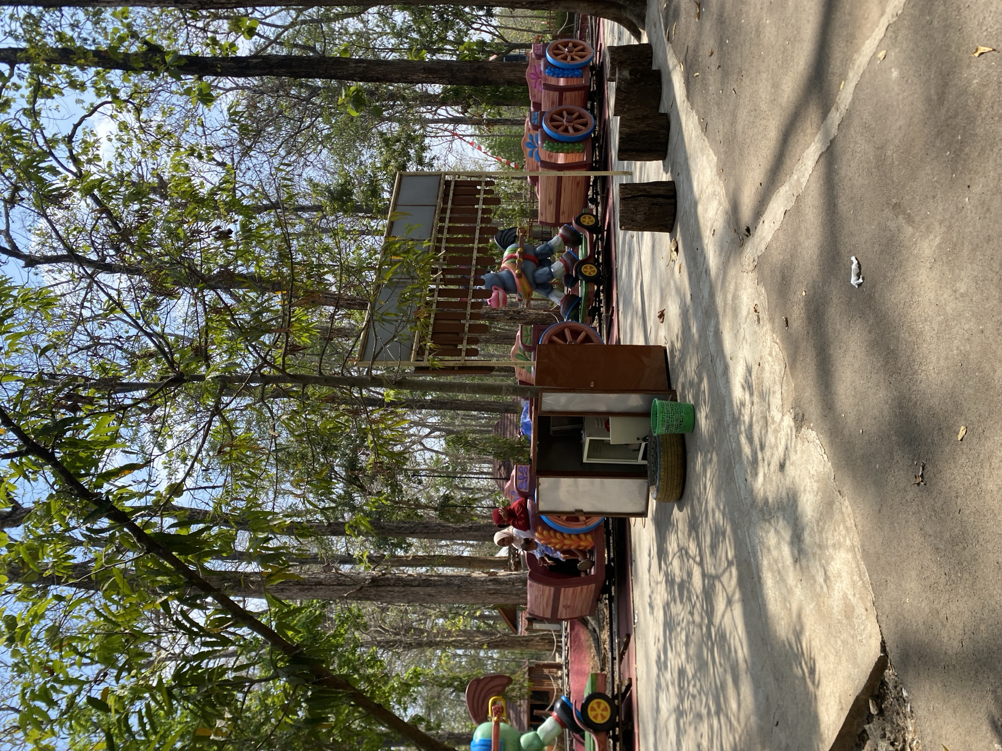 A sunny, outdoor scene at an amusement park shows a colorful train ride with wooden carriages designed to look like toy wagons. The train is pulled by a grey, horse-like figure with blue and pink accents. Several people, including children and adults, are seated in the carriages. The ride is set amidst tall trees with green foliage, casting dappled shadows on the concrete path in the foreground. There are also wooden stumps arranged on the path, a green trash can, and a discarded tire near a small kiosk. The overall atmosphere suggests a leisurely day at a recreational facility.