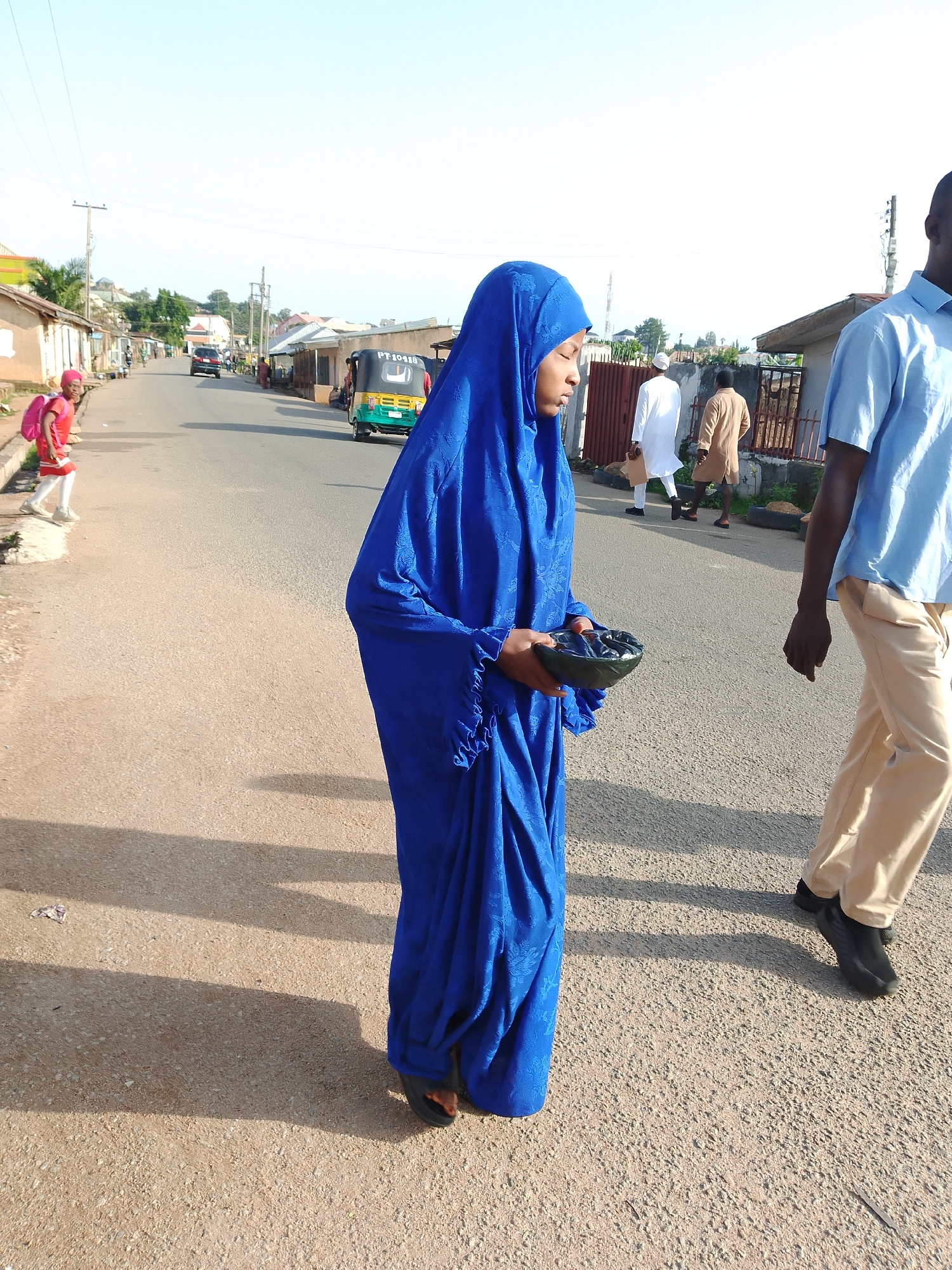 The image shows a street scene in Jos, Nigeria, on a clear, sunny day. A woman wearing a vibrant blue hijab and dress is in the foreground, holding a dark, shallow bowl in her hands. Her face is partially visible, with her eyes closed. To her right, a man in a light blue short-sleeved shirt and beige trousers walks away from the camera, his back mostly to the viewer. In the background, a young girl in a red outfit with a pink backpack is walking on the side of the road. Further down the street, several other people are visible, including two men in traditional attire, one in white and the other in brown. A yellow and green auto-rickshaw is also driving down the road. The street is paved, and there are buildings and shops on either side, with some trees and vegetation visible in the distance. The scene appears to be a typical urban environment in Jos, with everyday activities taking place.