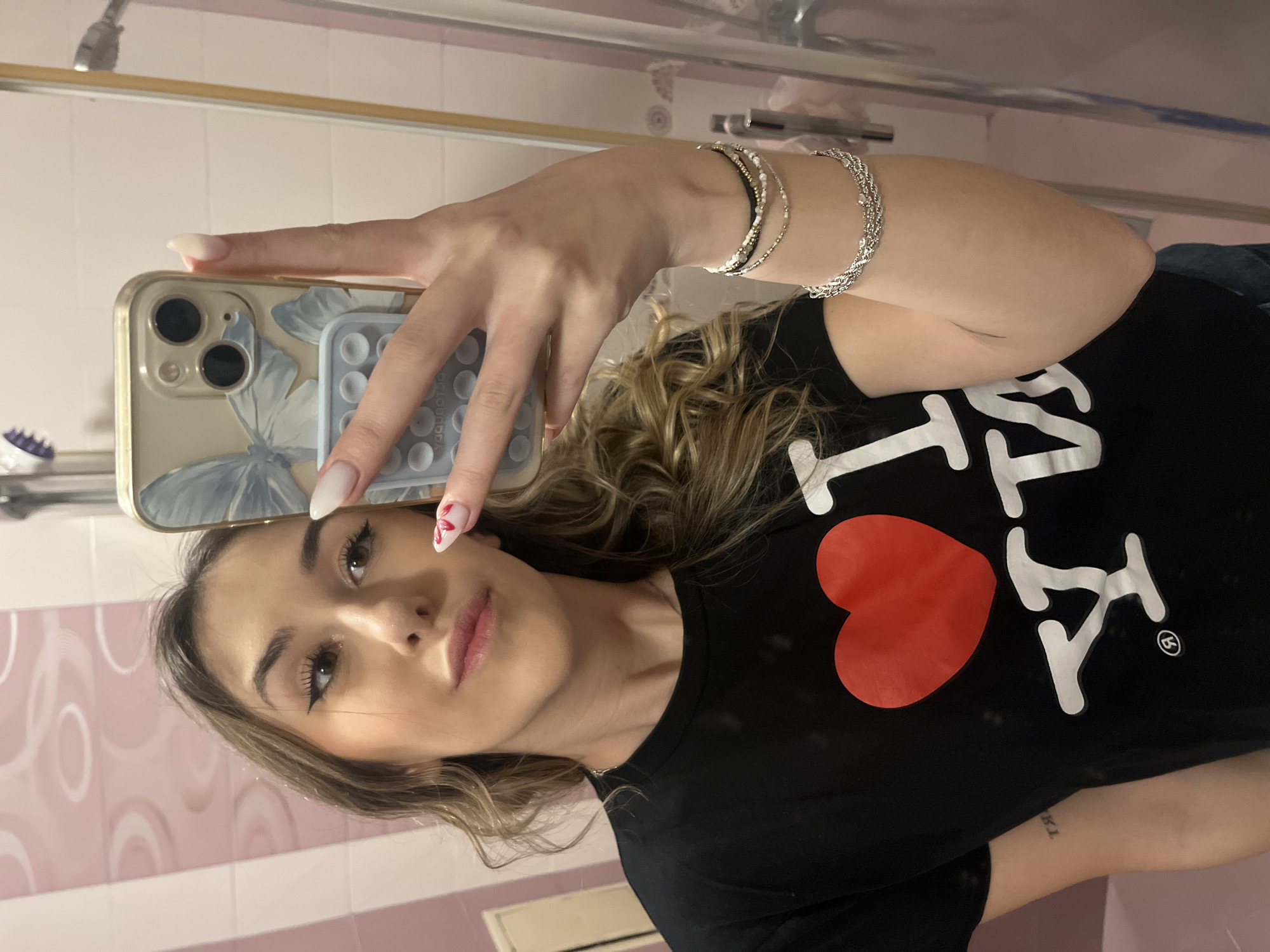 A fair-skinned woman with blonde hair is holding a smartphone in her left hand, taking a selfie in what appears to be a bathroom. The phone case has a butterfly design. She is wearing a black t-shirt with an "I (heart) NY" design. She is wearing multiple bracelets on her left wrist. The bathroom has pink and white tiled walls. The location cannot be determined as Sofia, Bulgaria without additional information.