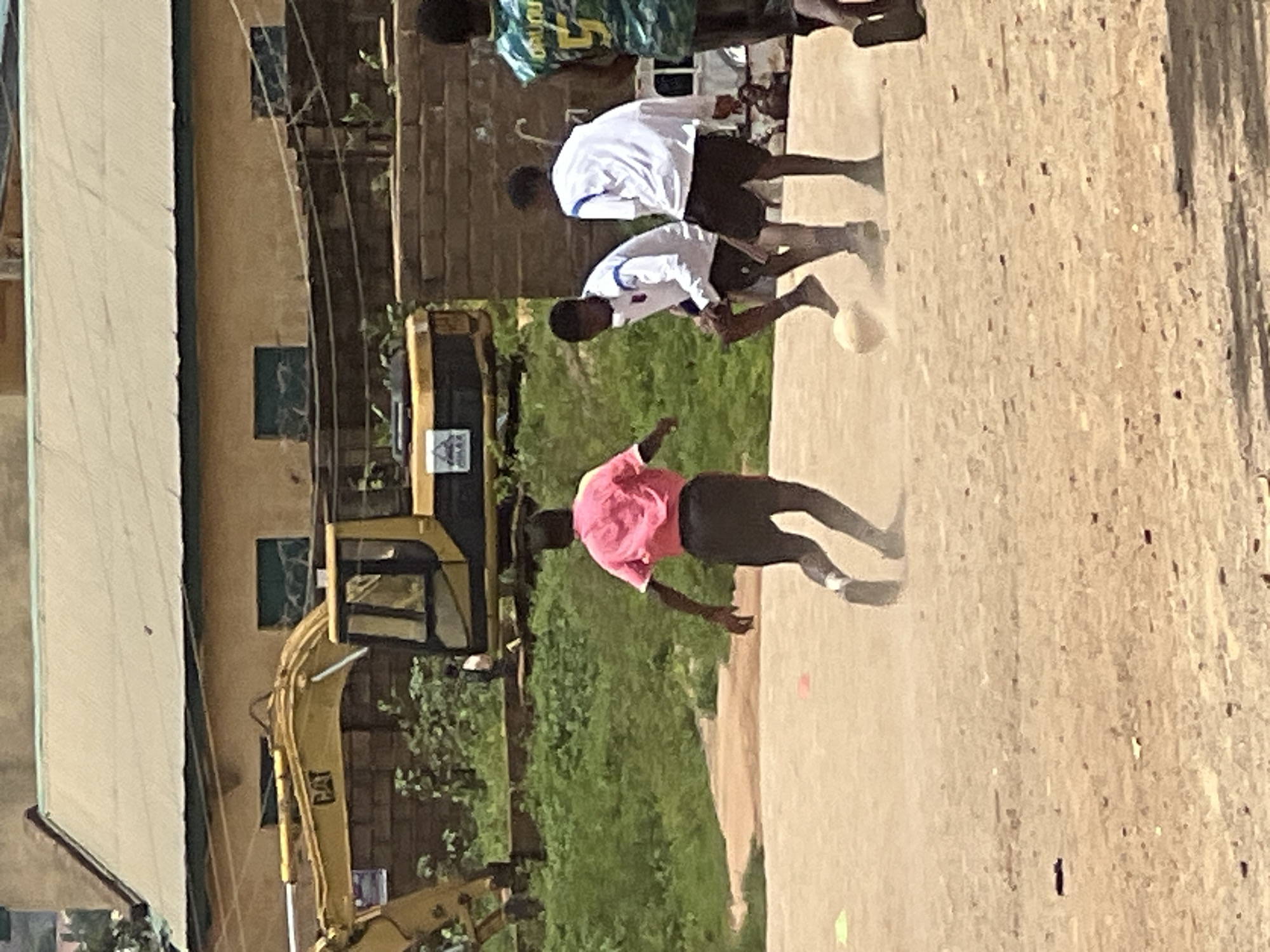 In the city of Jos, Nigeria, several young men are engaged in a game of soccer on a dusty, outdoor field. The foreground is dominated by the sandy ground, with visible dust kicked up by the players' movements. A soccer ball is in motion between two of the individuals. One player, wearing a pink t-shirt and dark shorts, is running away from the viewer, his back to the camera. Two other players in white shirts and dark shorts are in the center of the frame, actively playing the ball. In the background, a yellow excavator bearing the "CAT" logo is parked near a building with a tan-colored roof and yellow walls. Adjacent to the excavator is a rough brick wall. Green vegetation is visible behind the players and between the excavator and the brick wall. The overall scene suggests a casual outdoor sports activity taking place in an area with ongoing construction or development.