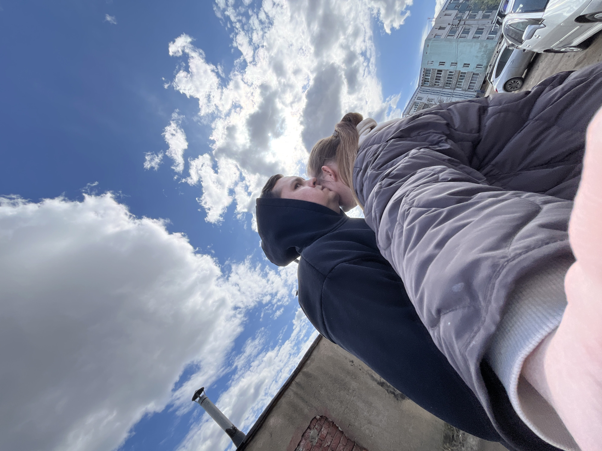 A photograph taken in Vysokovsk, Russia, shows a man and a woman kissing under a sky with scattered clouds. The man is wearing a dark hoodie, and the woman is wearing a quilted jacket. The background includes a building with multiple windows, parked cars, and the corner of a brick and concrete structure with a chimney. The photograph is taken at an upward angle, giving a distorted perspective.