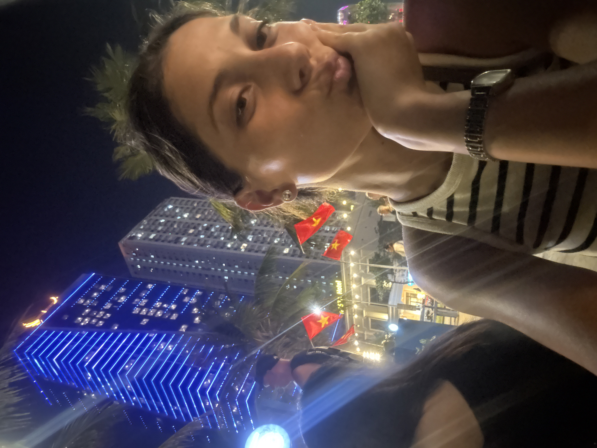 A tilted, low-angle nighttime shot captures a woman posing for a selfie in Đà Nẵng, Vietnam. The woman has light skin, dark hair, and is wearing a striped top and a watch. She is puckering her lips and holding her cheek with her hand.
In the background, illuminated buildings, including a tall one with blue lights, stand out against the dark sky. Vietnamese flags are displayed amidst palm trees and streetlights.