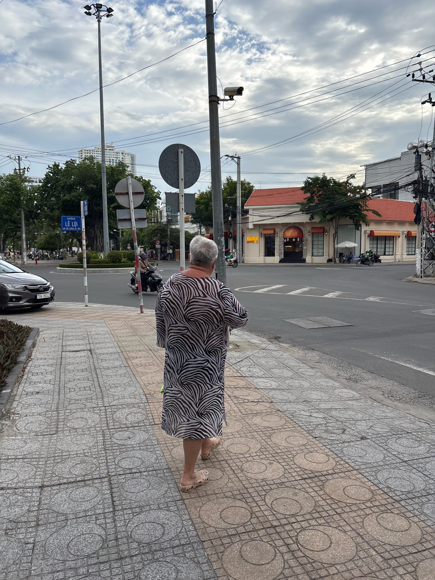 A woman with grey hair walks on a sidewalk in Nha Trang, Vietnam. She is wearing a black and white zebra-print dress and light-colored sandals. The sidewalk is paved with patterned tiles. In the background, there is a street with traffic, including a car and motorcycles. Buildings with red tile roofs are visible, as well as streetlights and power lines. The sky is cloudy. A street sign indicates "ĐƯỜNG LÊ LỢI".