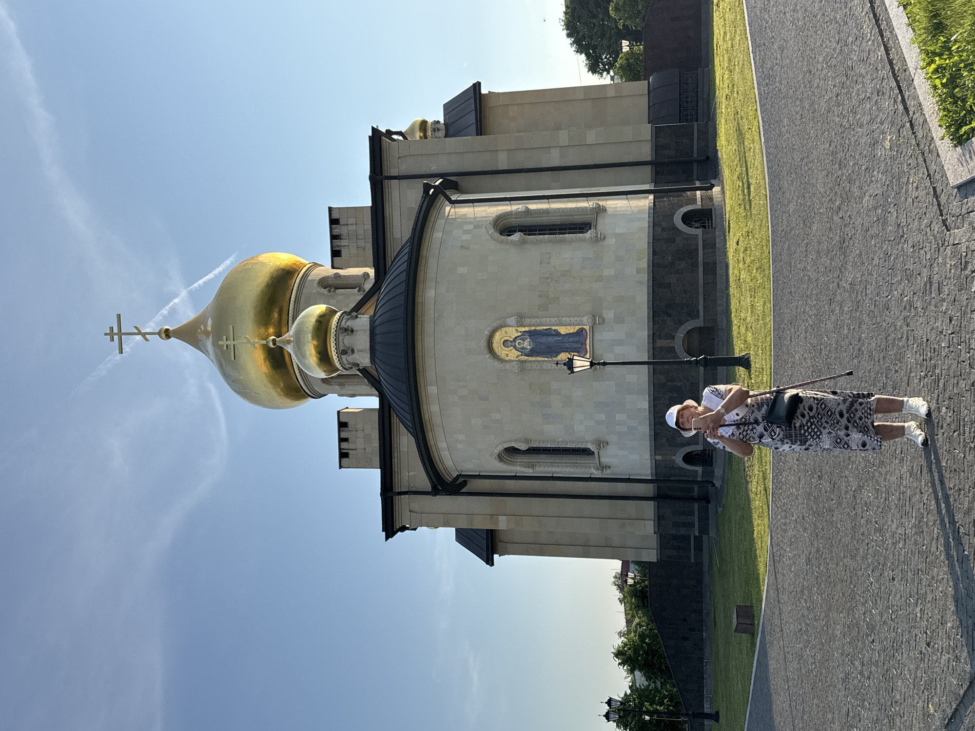 A woman stands on a cobblestone path in front of a large building with golden domes. She is wearing a white hat, a patterned dress, and white shoes, and is holding a cane and a phone. She appears to be taking a picture of herself or the building. The building, a church, has light-colored stone walls and a dark roof. The sky is blue with some wispy white clouds, suggesting it is daytime. The scene is well-lit, indicating it's likely sunny. The ground is a mix of cobblestones and green grass. The overall atmosphere is peaceful and serene. There is no visible text in the image. The location is identified as Kirishi, Russia.