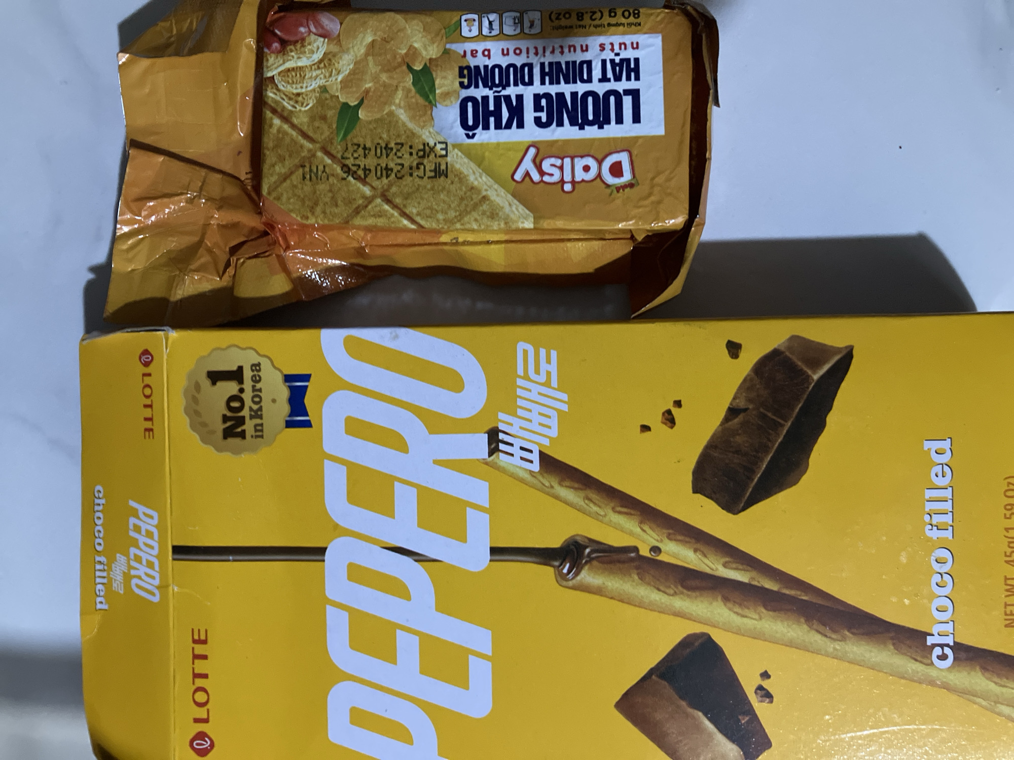 The image displays two snack food items on a white surface, casting shadows to the left. On the left is a partially visible yellow box for "PEPPERO" choco filled snacks, manufactured by LOTTE. The box features imagery of chocolate-dipped pretzel sticks and chocolate chunks. To the right is a partially unwrapped orange package of "Daisy" LƯƠNG KHÔ nuts nutrition bar, which indicates its net weight as 80g (2.8 oz). The context of Nha Trang, Vietnam is not discernible from the visual information provided in the image.