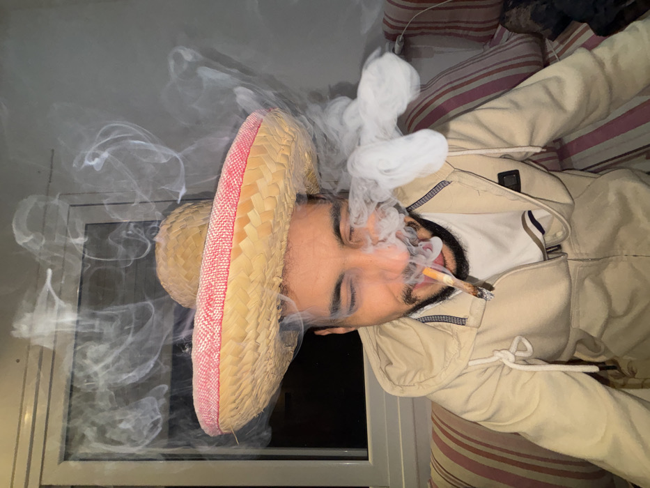 An image taken indoors in Martil, Morocco, depicts an adult male seated on a striped couch, exhaling a plume of white smoke. The subject wears a beige hooded jacket over a white t-shirt and a straw sombrero with a red and white patterned band. His eyes are closed, and a hand-rolled cigarette, glowing orange at its tip, is held between his lips. Dense white smoke emanates from his mouth and around his face, ascending towards the top of the frame. The background features a window or glass door on the left, reflecting a dark exterior, and a plain light-colored wall on the right. A striped cushion, primarily red and beige, is visible behind him.
