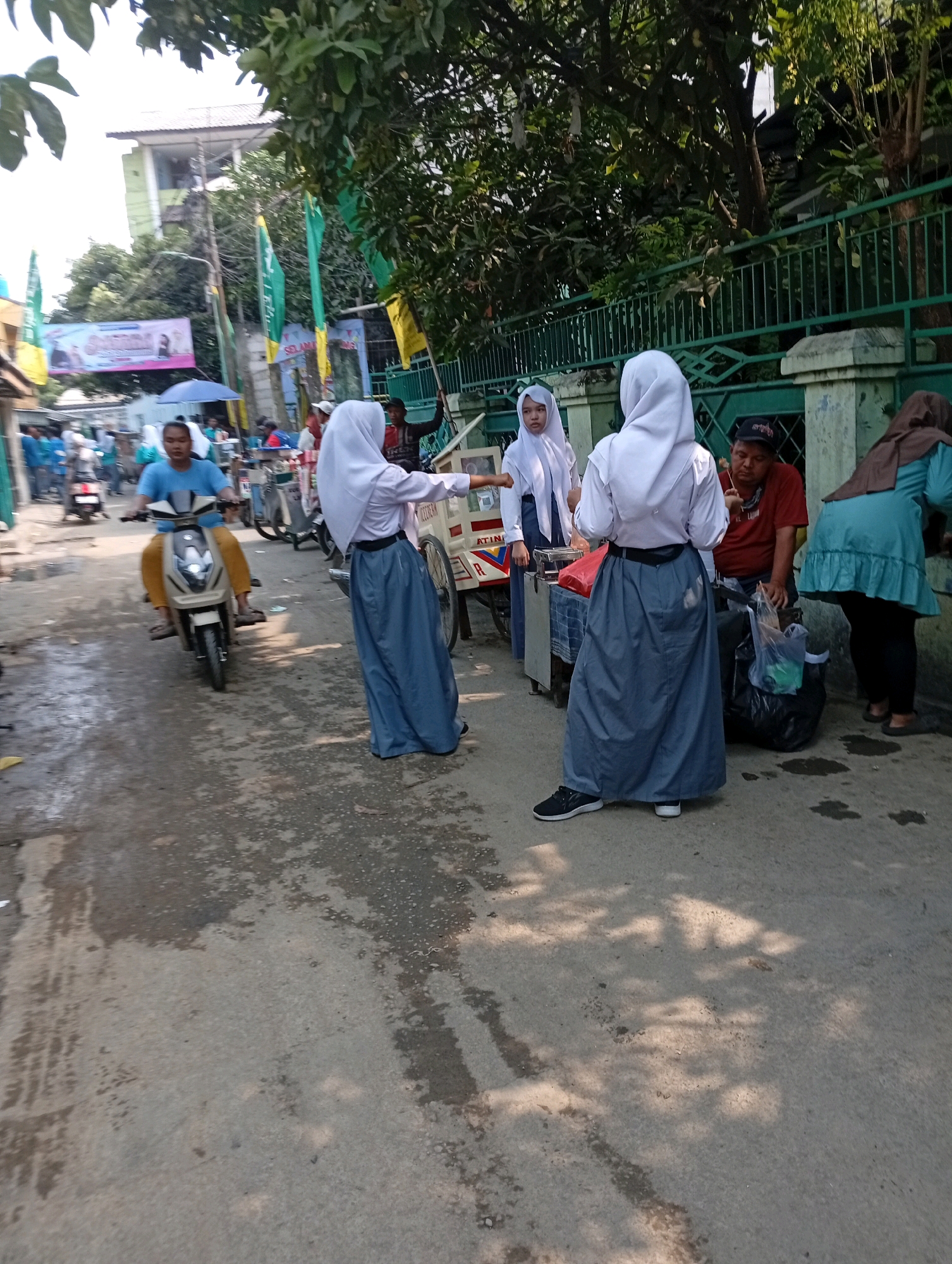 The image depicts a street scene in East Jakarta, Indonesia, on a sunny day. In the foreground, three young women in Islamic school uniforms, consisting of long gray skirts and white shirts, stand in the street. Two of them are facing away from the camera, and one is partially turned towards the viewer. They appear to be interacting with a vendor, a man wearing a red t-shirt and a black baseball cap, who is standing behind a food cart. The vendor is holding a skewer of food. To the right of the vendor, a woman in a turquoise abaya is also visible. In the midground, a young man wearing a blue t-shirt and yellow pants rides a scooter, moving from left to right. Several other people and motorbikes are present in the background, along with buildings and green fencing. Green and yellow flags are also visible, suggesting a local event or political gathering. The street is paved with asphalt and shows signs of being wet in some areas, with sunlight casting shadows.