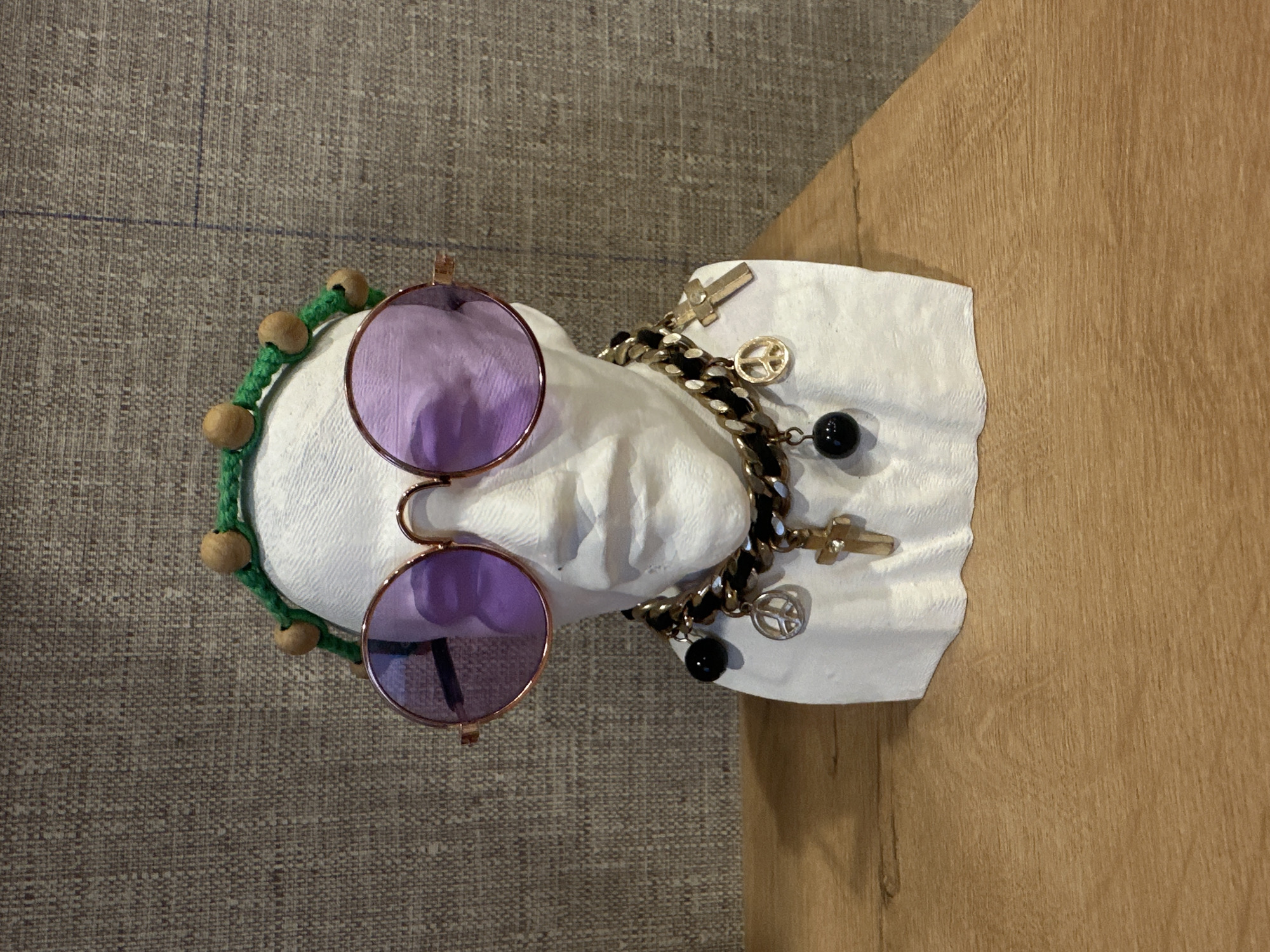 The image shows a pair of round, purple-tinted sunglasses resting on a white, crumpled cloth. A green beaded bracelet is also visible next to the sunglasses. A gold charm bracelet with religious symbols and black beads is draped over the cloth. The background consists of a gray textured surface and a light brown wooden surface. The location is not discernible from the provided image.