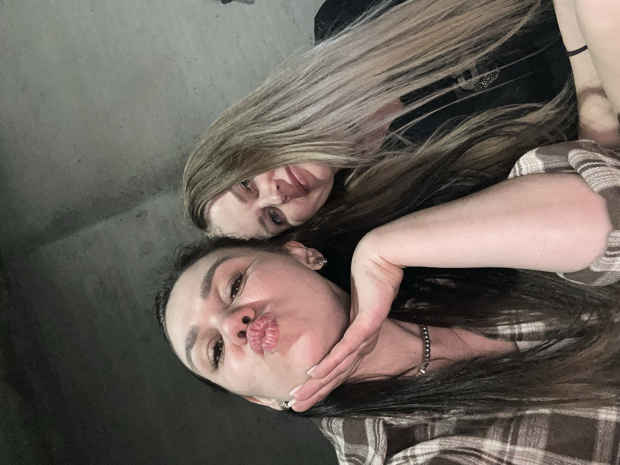 Two women are posing for a selfie. The woman in the foreground is pouting, while holding her hand under her chin. She has dark hair, dark eyebrows, and is wearing a plaid shirt. The woman in the background has blonde hair and is wearing a black top. The background appears to be a concrete wall. It is impossible to ascertain the location of the photo in relation to Noviy, Russia.