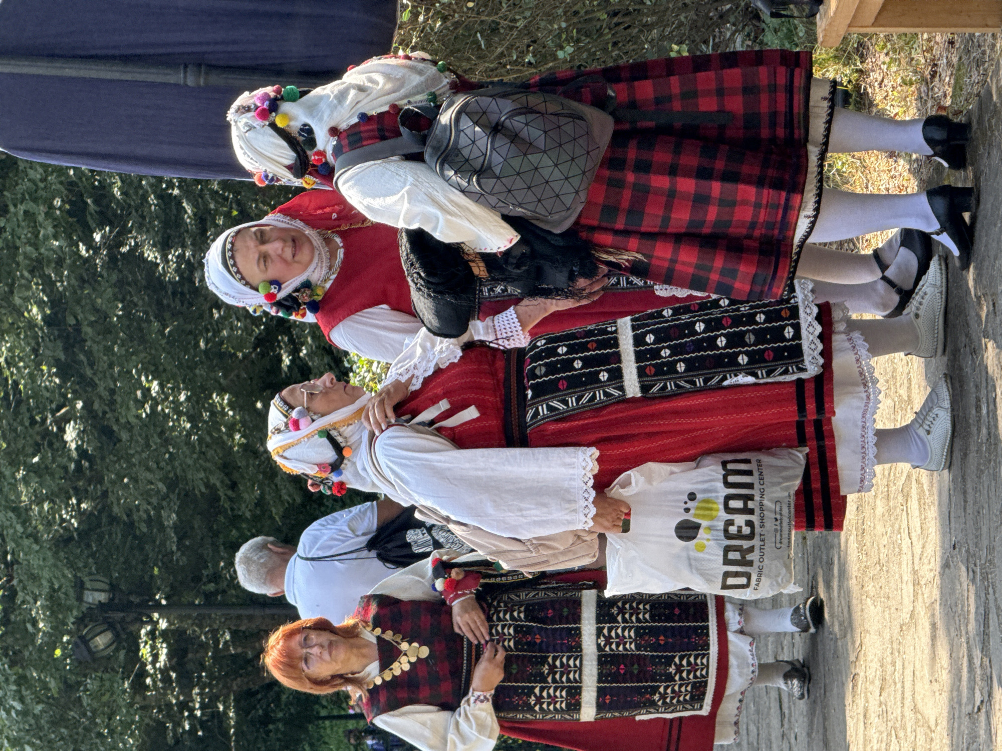 In the city of Ribaritsa, Bulgaria, a group of four individuals are standing outdoors on a paved surface, bathed in sunlight filtering through lush green foliage. The central focus is on three women dressed in traditional Bulgarian folk costumes. These costumes are vibrant and intricate, featuring red and black plaid skirts, white blouses with elaborate embroidery, and decorative headwear adorned with colorful pom-poms. One woman on the far left wears a red vest over a white blouse and a black and white patterned apron, accessorized with a gold necklace. She holds a white plastic shopping bag with "DREAM FABRIC OUTLET SHOPPING CENTER" printed on it. The woman next to her wears a similar red vest and apron, her headscarf featuring numerous colorful pompoms. The third woman, facing away from the camera, wears a red and black plaid dress and a white headscarf with pompoms, carrying a geometric patterned backpack. A fourth individual, a man with gray hair, stands behind them, partially obscured, wearing a white shirt. The background consists of dense green trees and a dark blue draped fabric, suggesting an outdoor event or festival. The overall atmosphere is lively and colorful, highlighting the cultural heritage of the region.