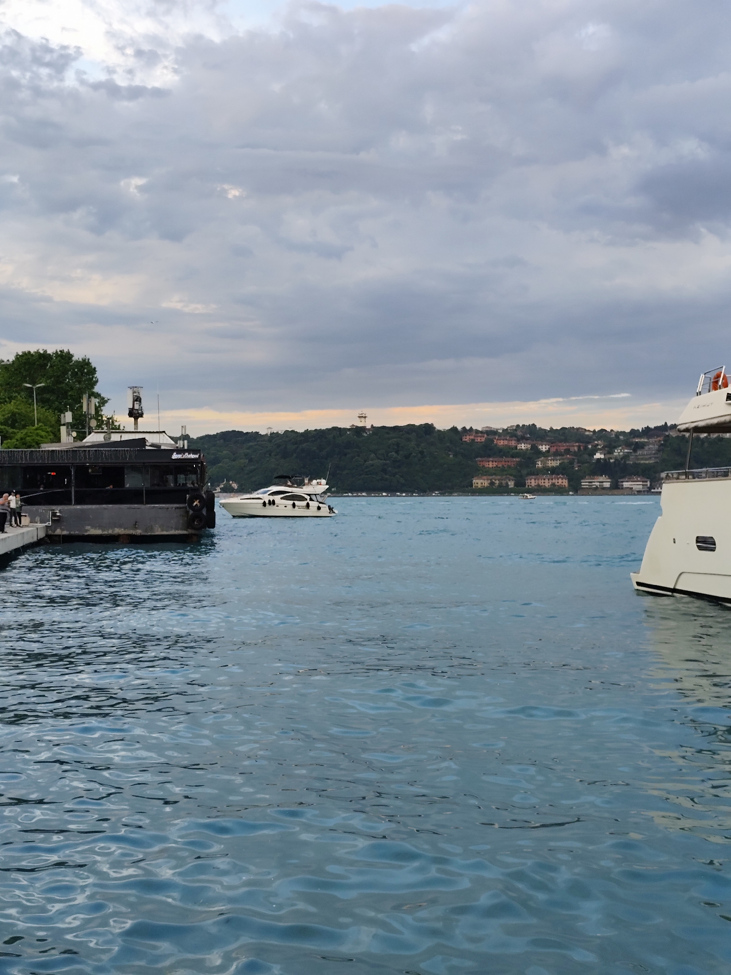 The scene depicts a body of water, likely the Bosphorus Strait, with a cloudy sky above. On the left, there is a dark-colored structure with people standing on a pier connected to it. To the right of this structure, a white yacht is visible on the water. In the center of the image, across the strait, there is a hillside with houses visible. On the right, a portion of a larger white yacht is seen. Given the location in Sarıyer, Turkey, the scene is likely a view of the Bosphorus, possibly near a dock or harbor area with maritime traffic and residential buildings along the shoreline. The time of day appears to be late afternoon or early evening, indicated by the sky.
