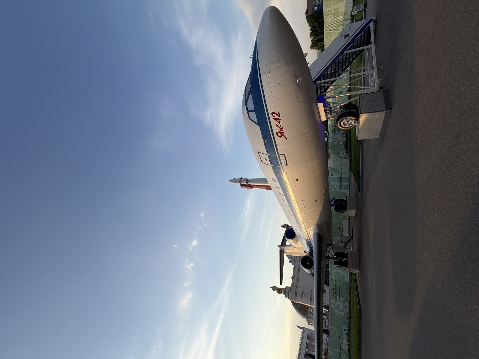 An aircraft bearing the inscription "ЯК-42" is on display at VDNH (Exhibition of Achievements of National Economy) in Moscow, Russia. The aircraft is silver and blue, with a boarding staircase attached. A rocket is visible in the background, along with architectural elements of the exhibition grounds. The sky is blue with wispy clouds. The scene is captured in daylight.