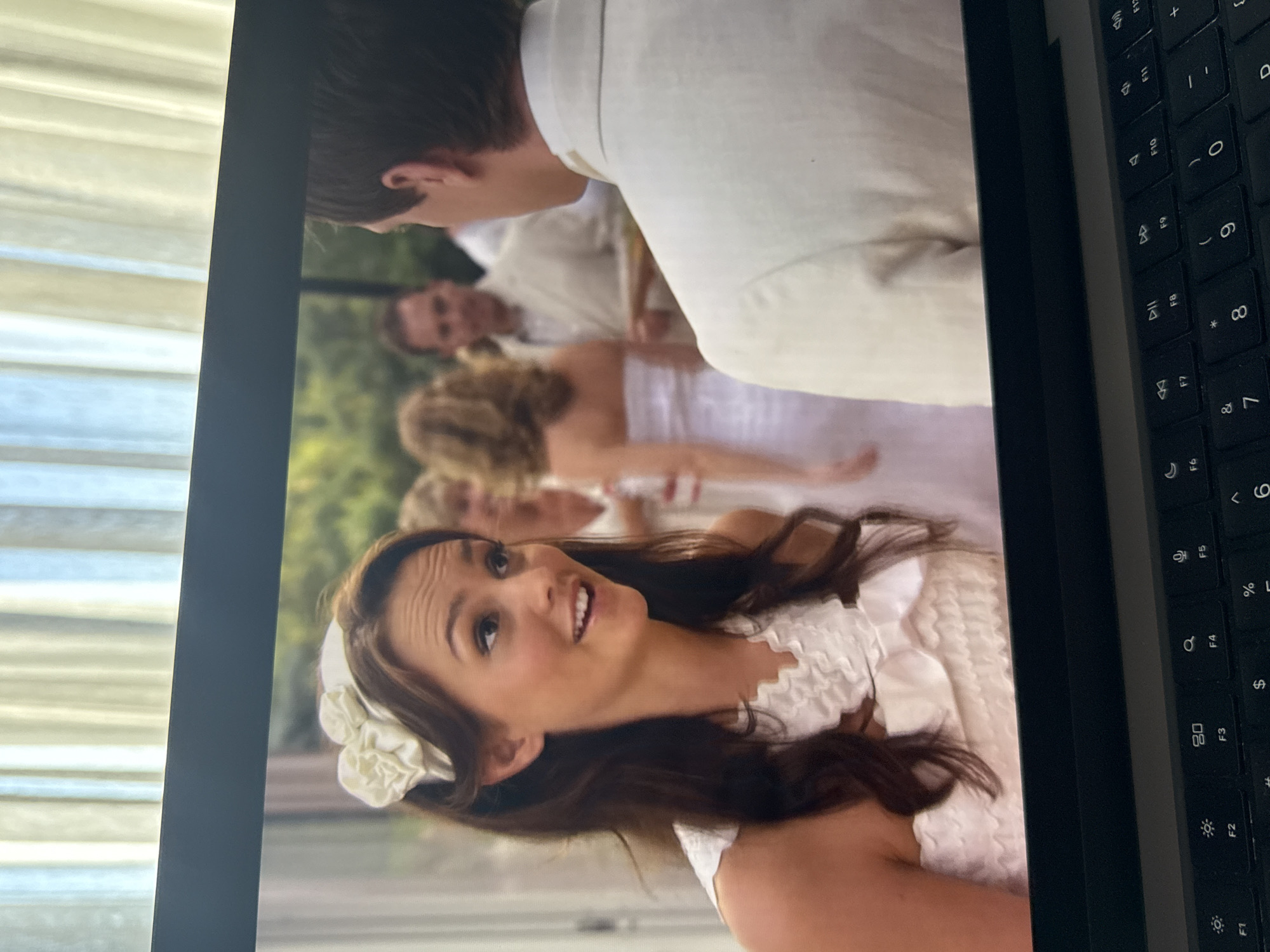 The image displays a laptop screen showing a scene with several people, taken from a slightly upward angle. In the foreground of the screen, a young woman with long, dark hair and a white floral accessory is visible. She wears a white, ruffled dress and looks upwards with an open-mouthed expression, suggesting surprise or amusement. Her face shows a slight smile. Behind her, partially blurred, are other individuals, including a man whose back is turned towards the viewer, wearing a light-colored suit, and other figures further in the background, some seated at a table. The setting within the screen appears to be an outdoor event, possibly a wedding or formal gathering, with greenery visible in the background, implying a garden or park setting during daytime.
The physical environment where the photograph was taken shows a dark-colored laptop with a keyboard visible on the right side of the frame, displaying function keys (F1 through F12) and number/symbol keys (e.g., F5, F6, 7, 8, 9, 0, %, &, *). On the left side, light-colored horizontal blinds or curtains are partially visible, suggesting an indoor setting with natural light coming through a window. The overall image captures a moment from a video or picture on a screen, within a room in Yerevan, Armenia.