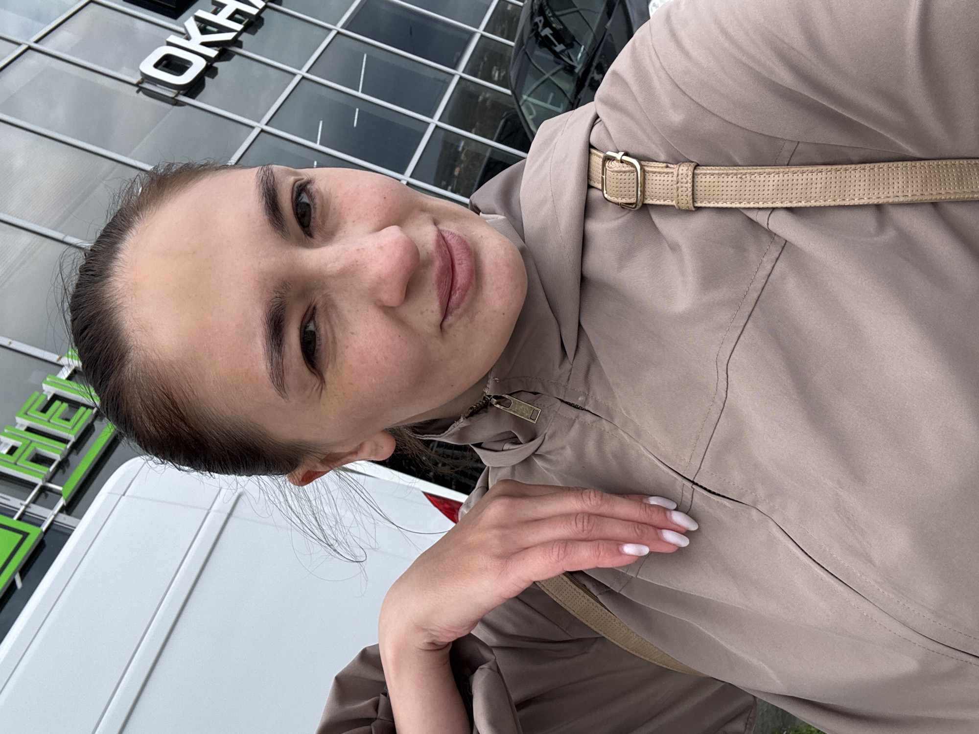 A woman in a light brown jacket takes a selfie outside a building in Sochi, Russia. The building has a sign with the word "OKHA" visible. A white vehicle is partially visible on the left side of the frame. The woman has dark hair pulled back, fair skin, and light pink lipstick. Her left hand is visible, with white polished nails. The jacket has a hood and a strap with a buckle. The lighting suggests an overcast day.