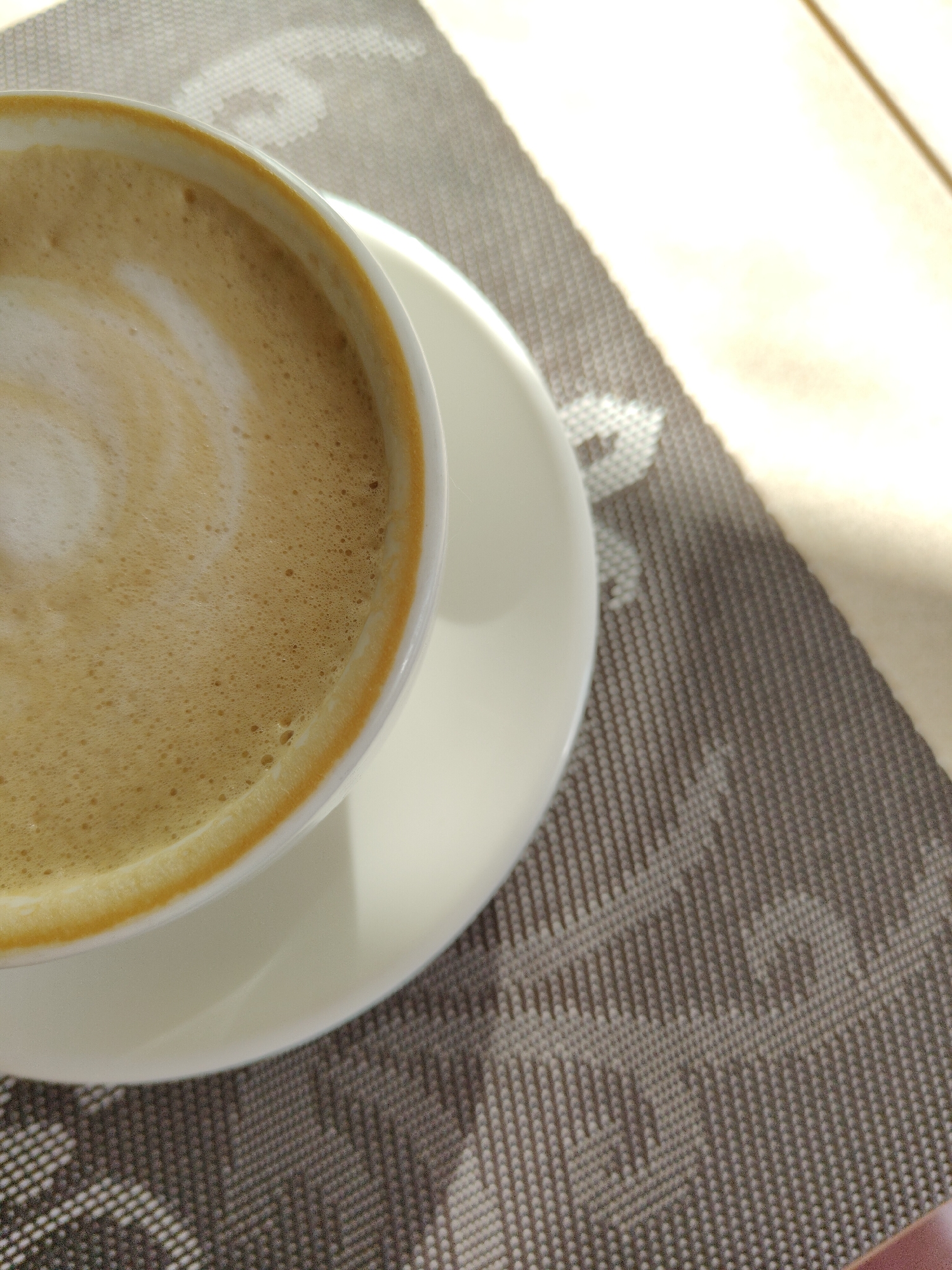 A close-up shot shows a cup of coffee with frothy foam on a white saucer, sitting on a patterned placemat. The lighting is bright, with a strong light source coming from the right side of the frame. The scene is likely indoors, but the specific location and city are unknown. There are no people visible in the image. The focus is on the coffee and the immediate surface it sits on. The environment appears clean and orderly.