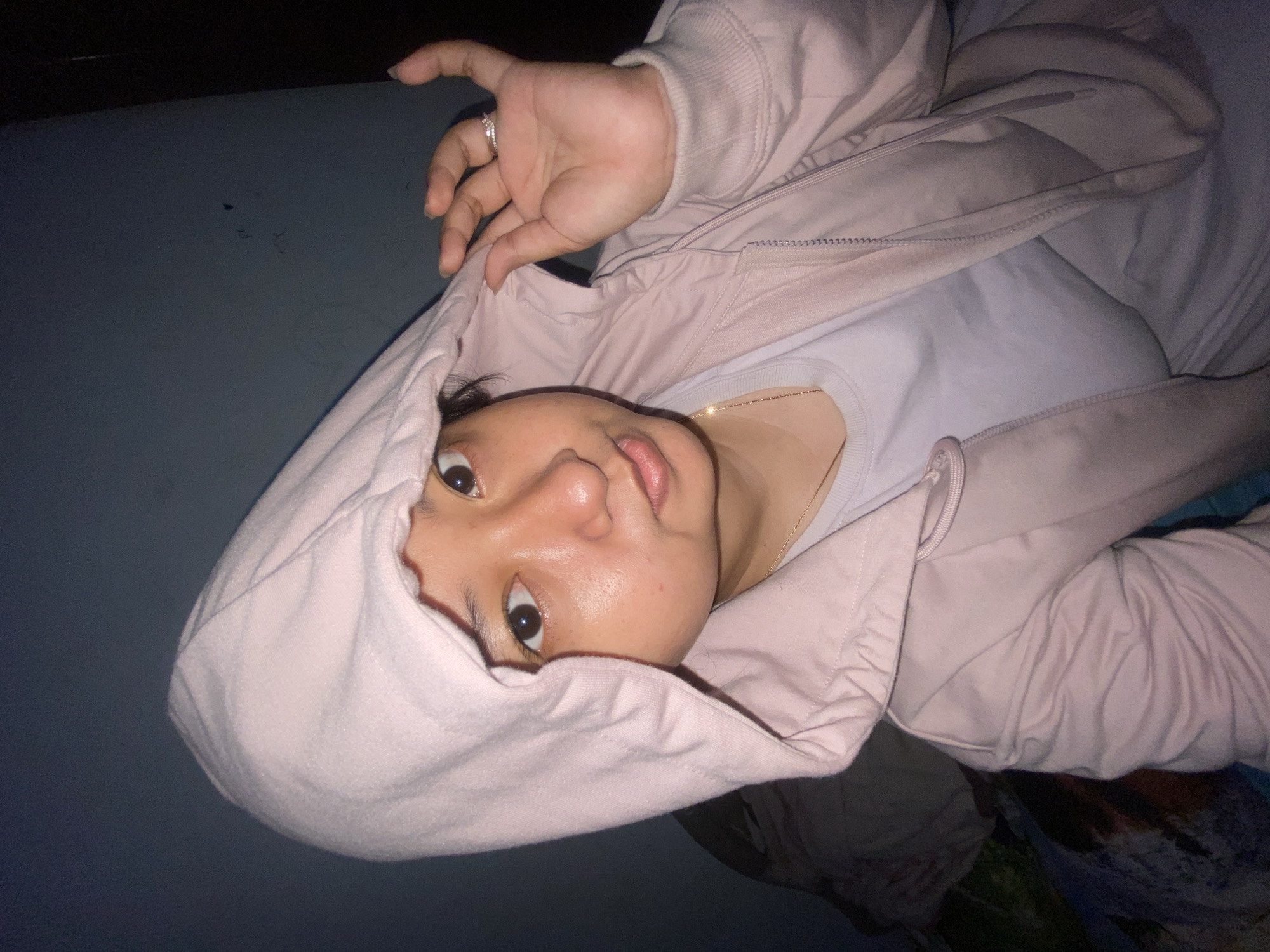 A young woman is shown from the chest up, pulling the hood of her light pink jacket over her head. She is wearing a light purple t-shirt underneath the jacket. She has dark hair, dark eyes, and a light complexion. She is wearing a ring on her right index finger. The background appears to be a dark blue wall. The lighting is dim, suggesting the photo was taken indoors at night. There are no discernible landmarks or environmental features that would indicate a specific location within Gelam, Indonesia.