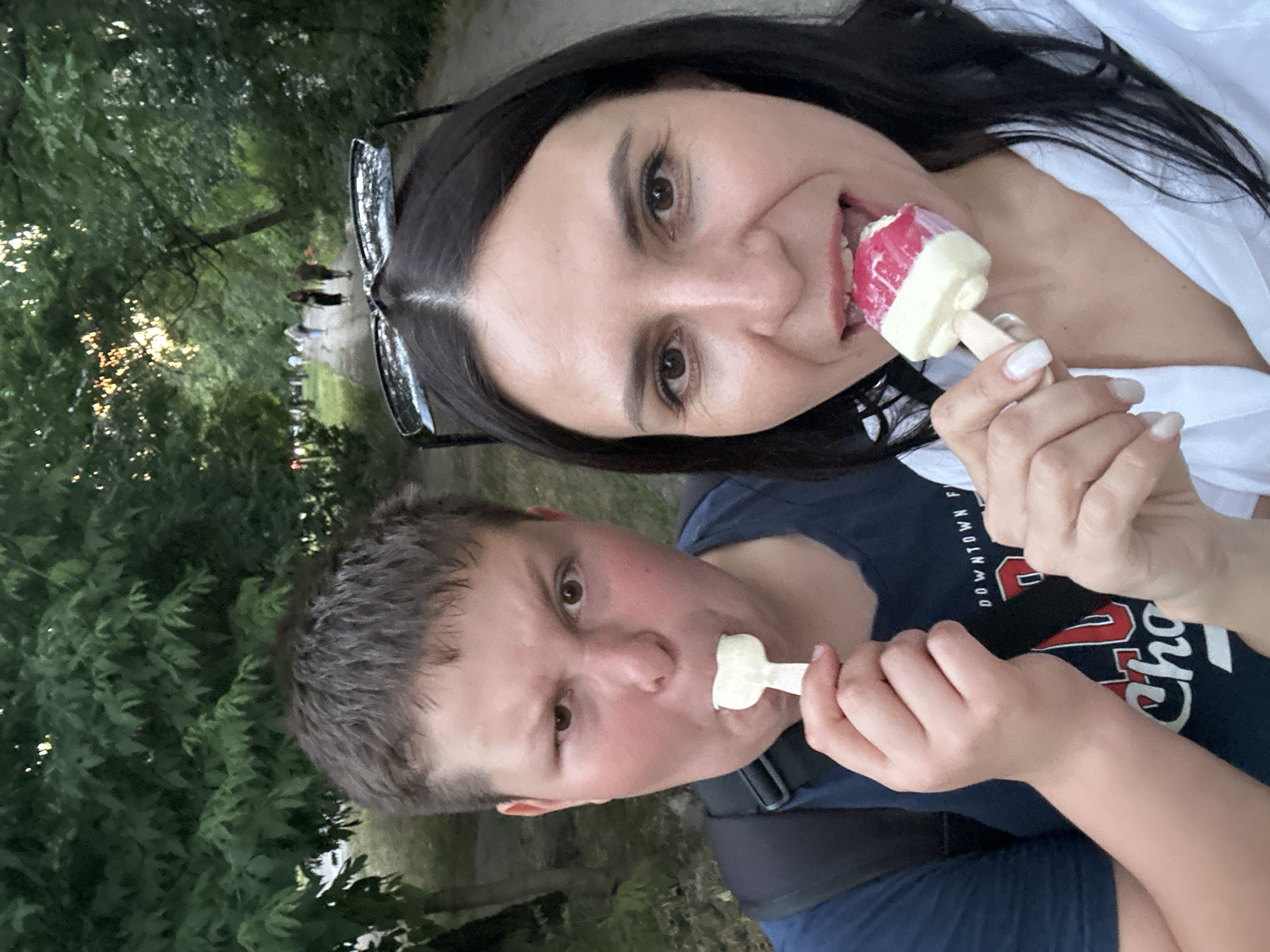 The media file shows a close-up, selfie-style shot of two people, likely a woman and a younger person, in an outdoor setting. They are both holding and eating ice cream popsicles.
The woman in the foreground has dark hair and is wearing glasses perched on her head. She is smiling and has the pink and white popsicle in her mouth. Her nails are painted a light color. The younger person, in front of her, has short brown hair and is looking directly at the camera with a slight grimace, holding a white popsicle to their mouth. They are wearing a dark blue t-shirt with some text and a backpack strap visible.
The background consists of lush green foliage and trees, suggesting a park or wooded area. Sunlight filters through the leaves, indicating it is daytime, likely late afternoon or early evening. The ground appears to be a path.
The overall mood seems casual and relaxed, with both individuals enjoying their ice cream. The scene is set in Odesa, Ukraine.
There is some visible text on the younger person's t-shirt, which reads "DOWNTOWN" and part of "CHO".