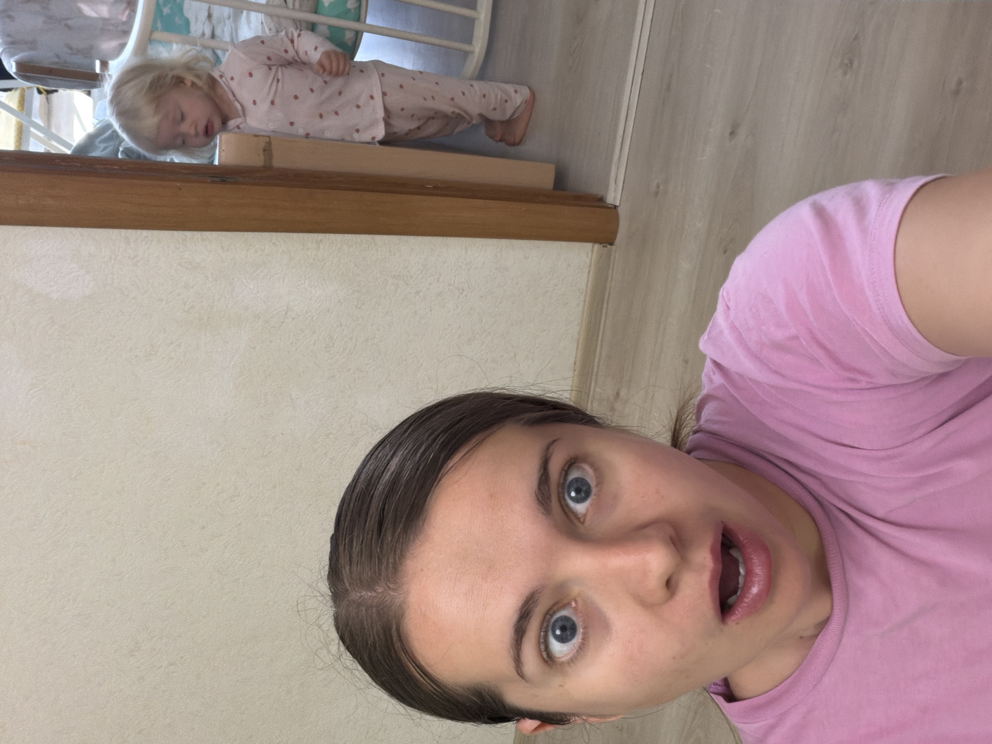 The image captures an indoor scene featuring an adult woman in the foreground and a sleeping child in the background. The entire photo is rotated clockwise, causing the subjects to appear tilted.
The woman, located in the right and bottom portions of the frame, is the primary subject. She has dark hair pulled back, fair skin, and striking wide blue eyes expressing surprise or shock, with her mouth agape. She is wearing a plain pink t-shirt. Her pose suggests she is taking a selfie.
In the upper left background, a young child with blonde hair is seen soundly asleep. The child wears light pink pajamas adorned with a subtle strawberry pattern. Their head rests face down on a horizontal, light brown wooden beam or railing. The child's bare feet are also visible further along this beam, indicating they are lying horizontally across this structure. A white crib or playpen railing is partially visible behind the child.
The setting appears to be a typical indoor domestic environment. The floor, visible on the right and top, is a light wooden laminate. The walls, on the left and bottom, are a light, textured beige or off-white. A darker wooden trim runs horizontally through the middle, separating the foreground from the background and serving as the surface the child is sleeping on.
The activity captured is the woman's surprised reaction, possibly to the child's unexpected sleeping position. The child, conversely, is deeply and peacefully asleep. The woman's wide eyes and open mouth clearly convey her astonishment, while the child's closed eyes and relaxed posture denote a profound slumber. The room is brightly lit by ambient indoor light. The photograph's slightly low and tilted angle adds to its spontaneous feel. There is no visible text in the image. Although the location context is Sochi, Russia, no specific visual cues within the image denote this geographical location, showing a generic residential interior.