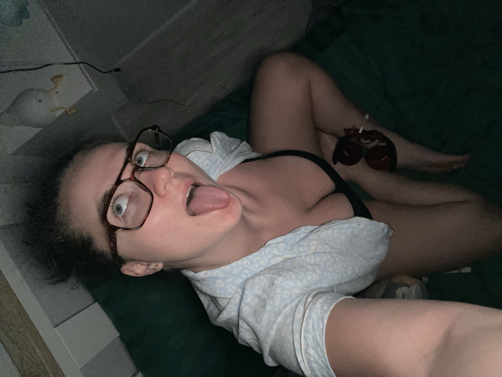A woman with glasses is lying on her back on a green surface, possibly a bed, in a dimly lit room. She is wearing a light-colored, patterned top. Her mouth is open and her tongue is sticking out. A pair of red headphones is resting on her leg. A white duck-shaped object is visible in the background, near a white wall. The location of the image is unknown; the specified city is irrelevant.