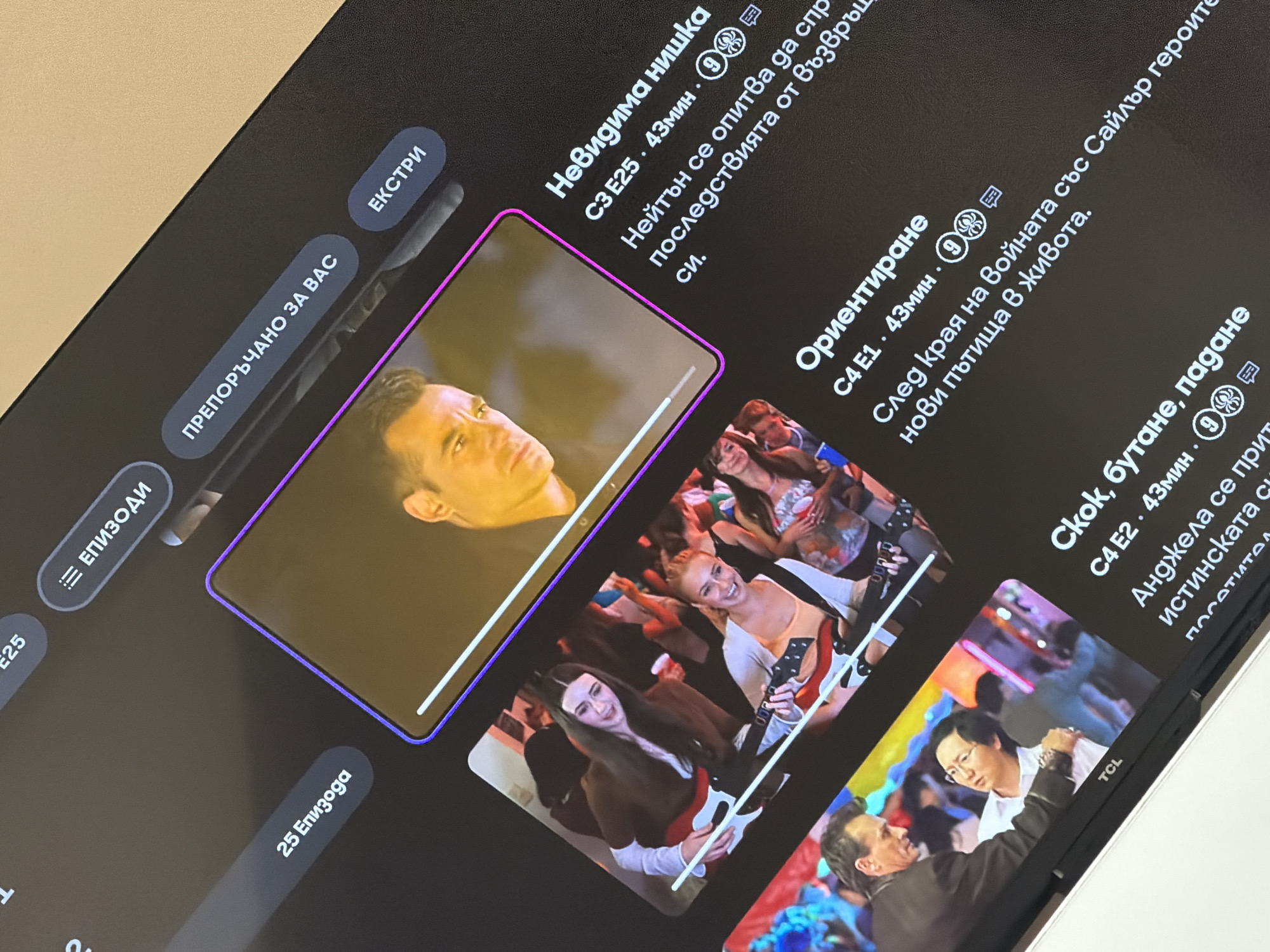 Here is a forensic description of the image:
The image shows a close-up of a screen displaying streaming content. The screen is dark with several video thumbnails arranged horizontally. Text accompanies each thumbnail, indicating show titles and episode information. The thumbnails feature a variety of scenes with multiple people. The screen also has menu buttons labeled "EPISODI" (Episodes), "ПРЕПОРЪЧАНО ЗА ВАС" (Recommended For You), and "EKCTРИ" (Extras). The bottom corner of the screen shows the brand "TCL." The language on the screen is Bulgarian. There is no identifiable location.