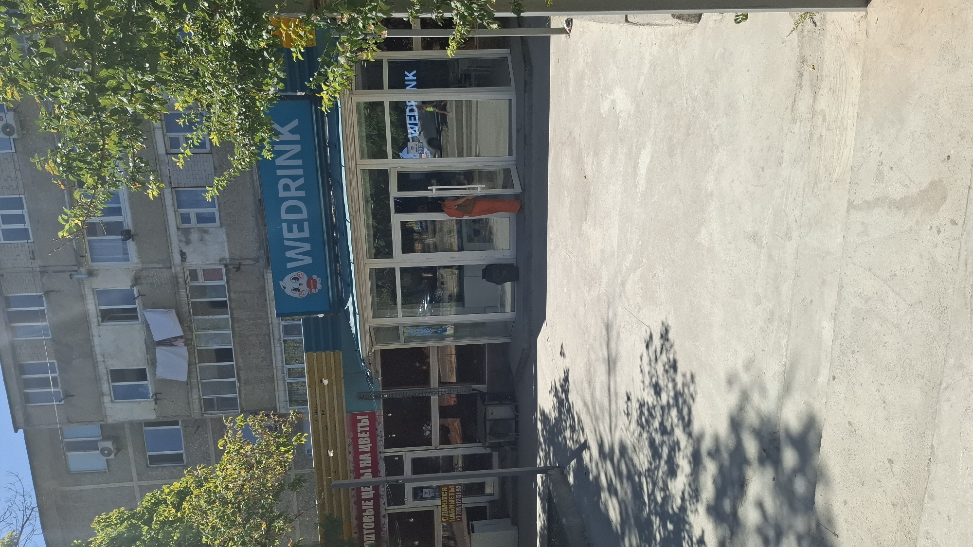 The image depicts the exterior of commercial establishments in Shymkent, Kazakhstan. In the foreground, a paved concrete area is visible with dappled sunlight casting shadows from trees. Behind this, a storefront with large glass windows and doors is prominent. The signage above this storefront reads "WEDRINK" in large blue letters, accompanied by a cartoon character logo. To the left of the "WEDRINK" establishment, another shop with a sign that includes Russian text, possibly "ОПТОВЫЕ ЦЕНЫ НА ЦВЕТЫ" (Wholesale prices for flowers), is visible. Further left, a sign that says "СДАЮТСЯ КАБИНЕТЫ" (Offices for rent) with a phone number is also present. A person wearing an orange garment stands near the entrance of the "WEDRINK" store. Behind the storefronts, a multi-story residential building with numerous windows and balconies, where laundry is hanging on some balconies, is visible. The overall scene suggests a commercial street or area within the city.